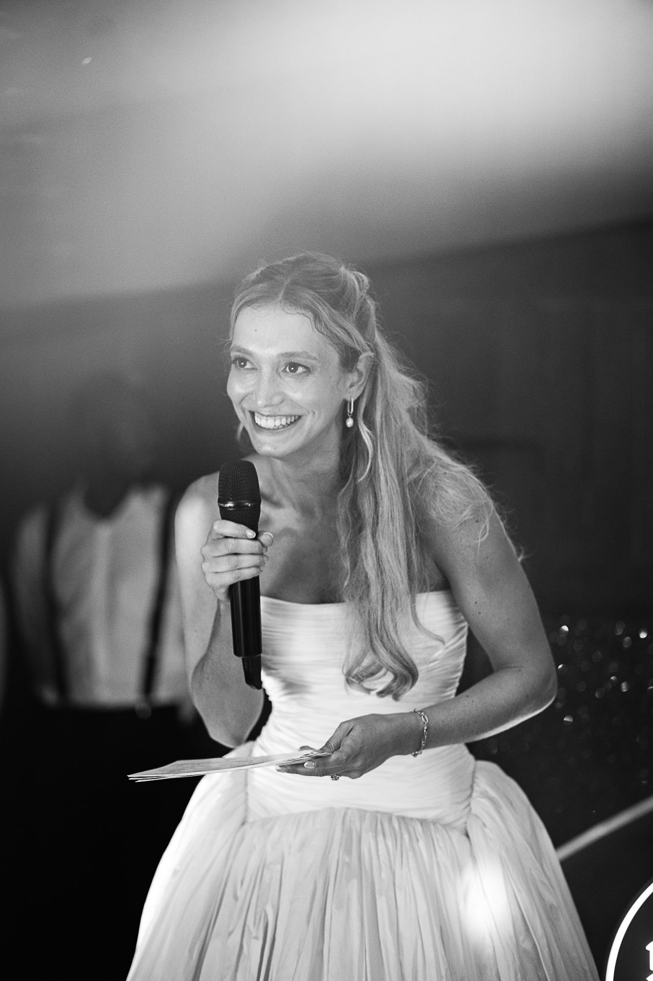 Smiling bride in white strapless gown holds microphone while giving speech at wedding reception, black and white photo.