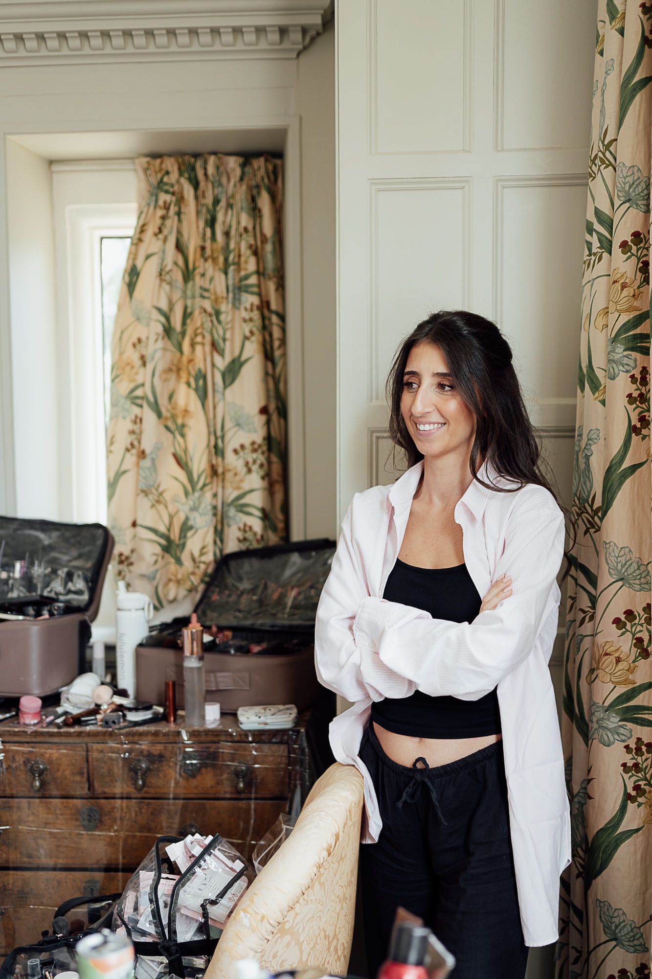 A smiling woman with dark hair stands arms crossed in a room with floral curtains and a makeup artist's kit.