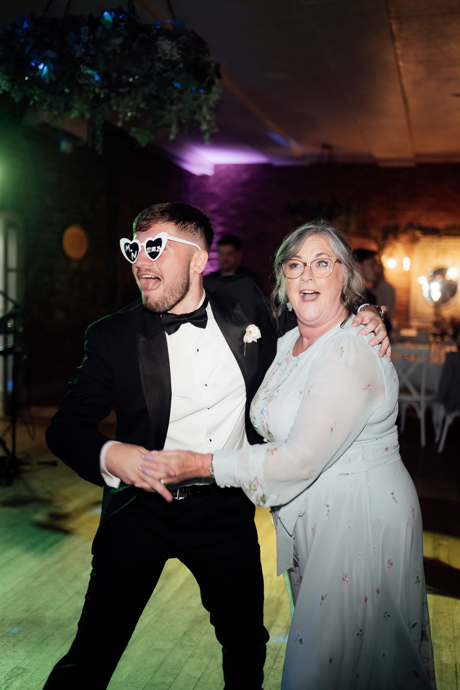 Two wedding guests dance enthusiastically on a dance floor with one wearing sunglasses and a bow tie.