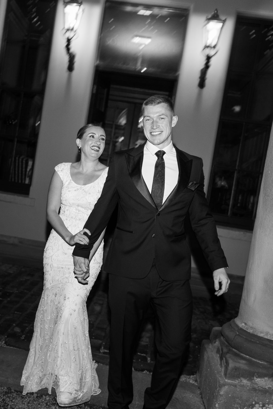 A laughing couple dressed in formal wedding attire walk together during an evening celebration.