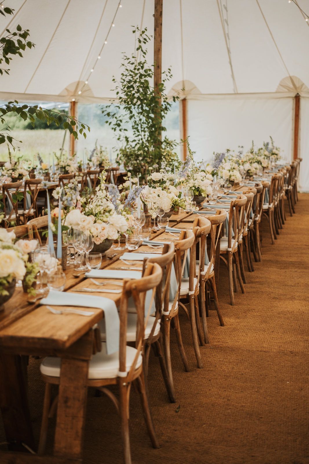 Long wooden farm tables and cross-back chairs create elegant rows inside a white tent for an outdoor wedding reception.