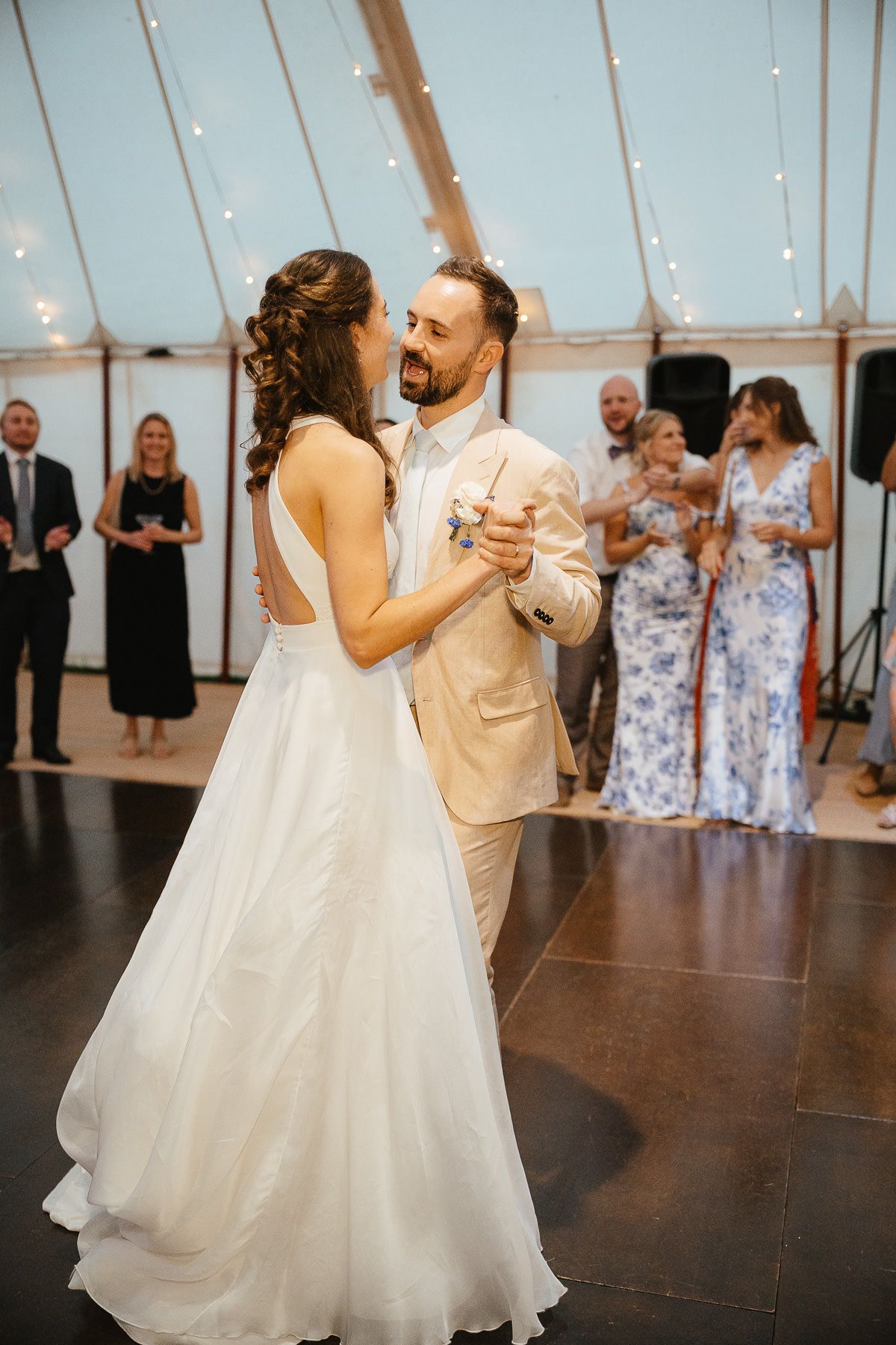 A couple dances together on a dark floor while guests watch during a wedding reception in a tent with string lights.