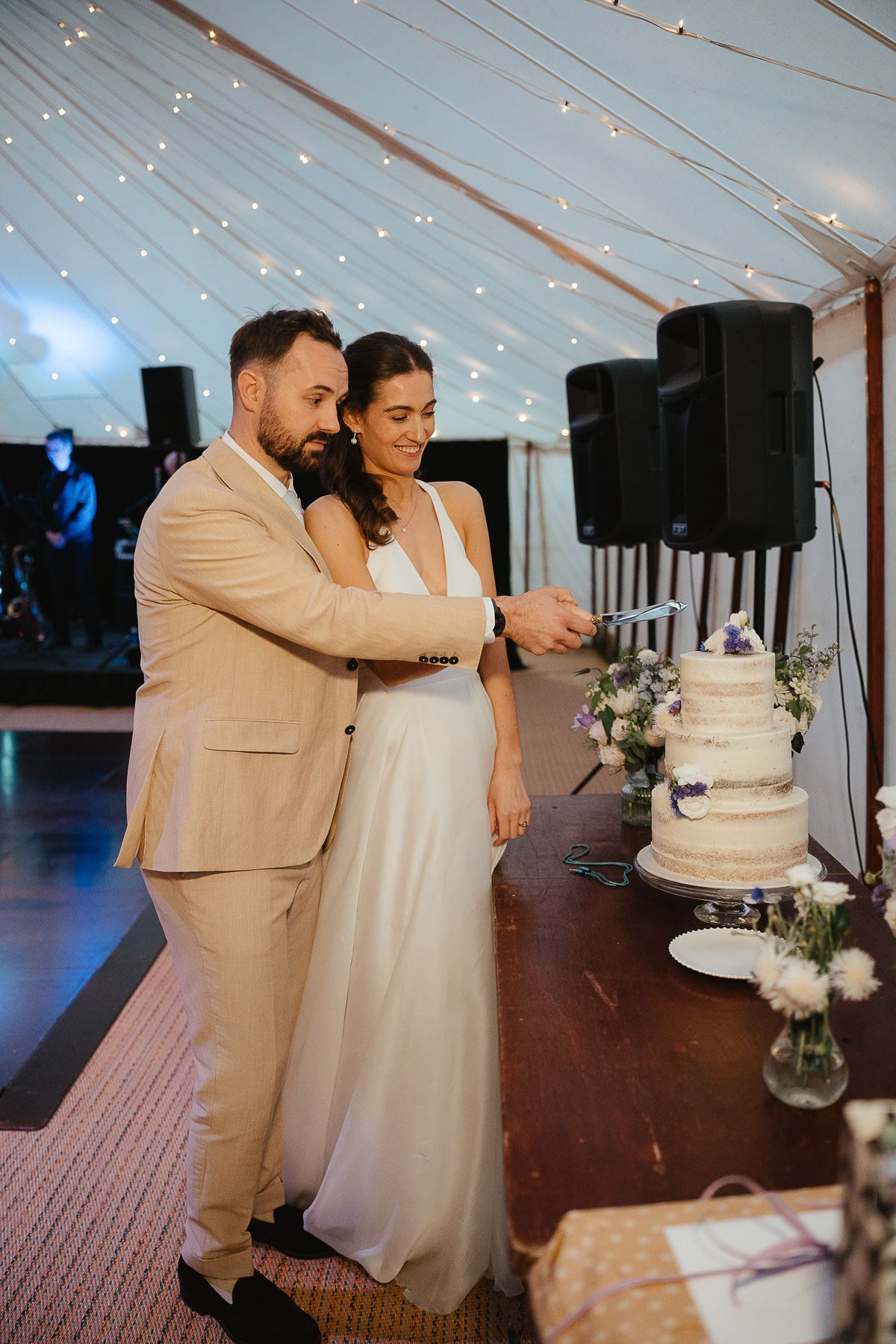 Couple cuts their elegant white wedding cake together under a twinkling tent at twilight reception.