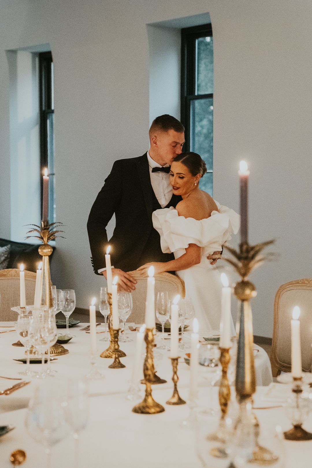 A romantic wedding reception table set with glowing candles in brass holders as a couple shares an intimate moment.