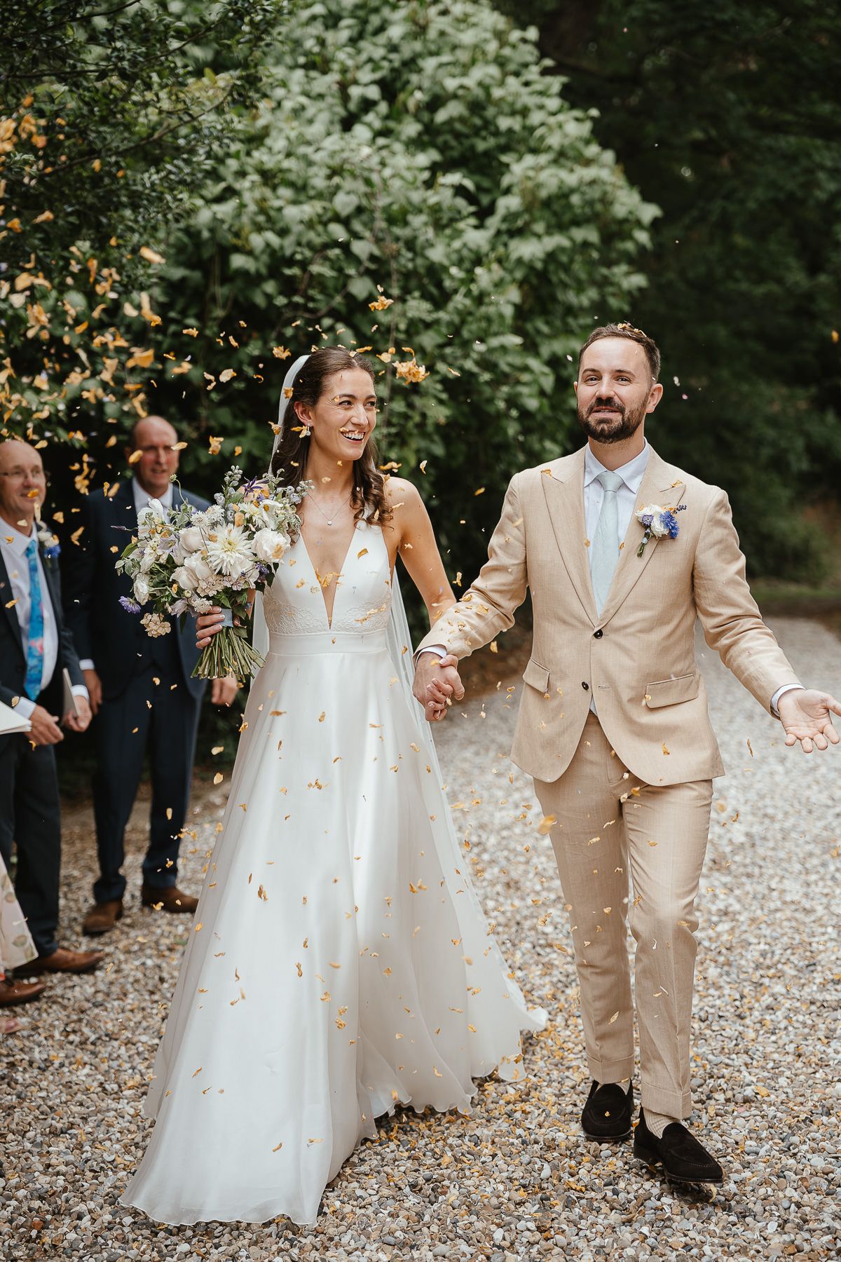 Wedding couple and guests walk down path while flower petals fall during outdoor garden celebration.