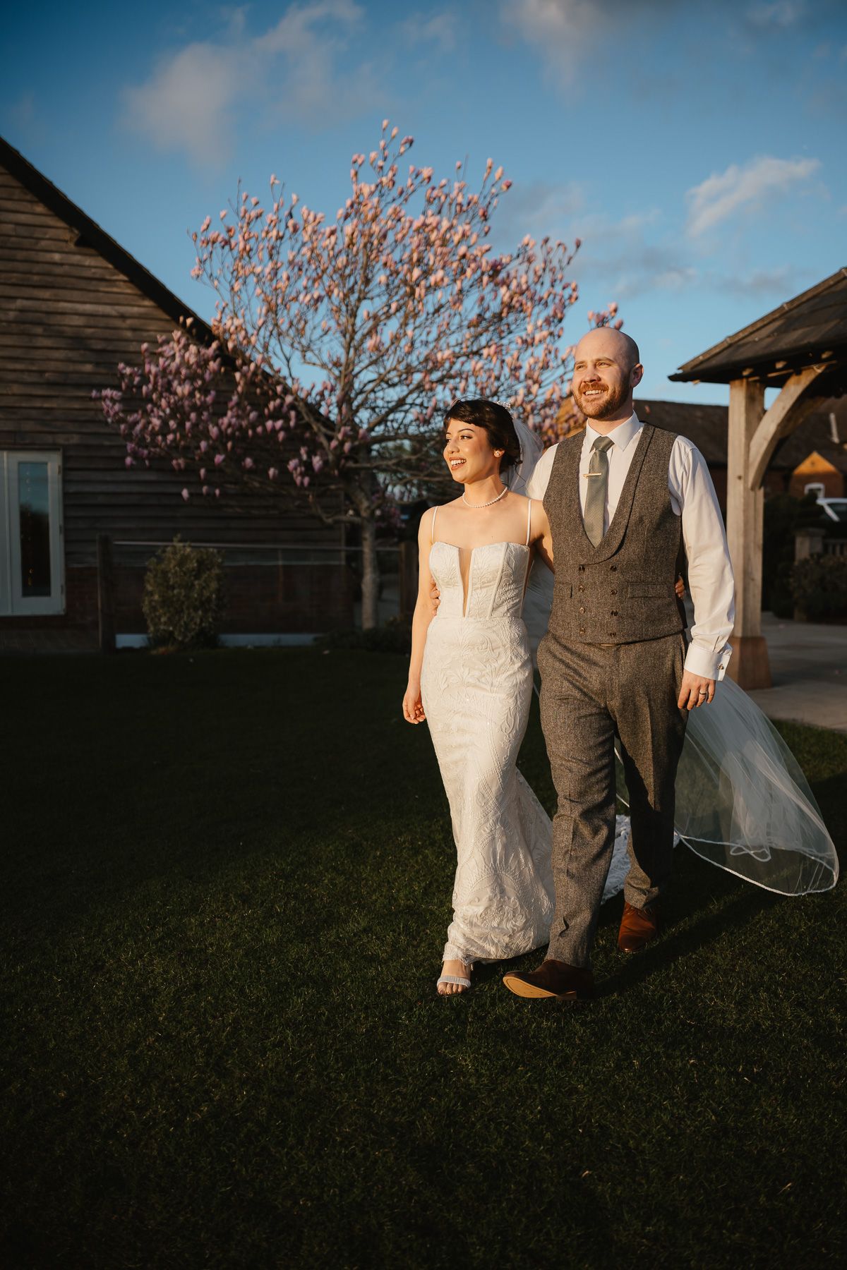A couple walks together on their wedding day in front of a rustic barn with a blooming tree in the background at sunset.