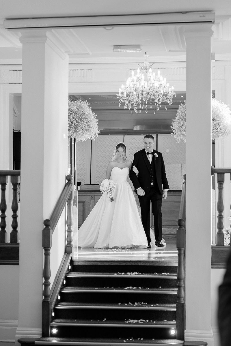 A sequence of black and white photos showing a bride and groom walking down elegant stairs with chandeliers overhead.