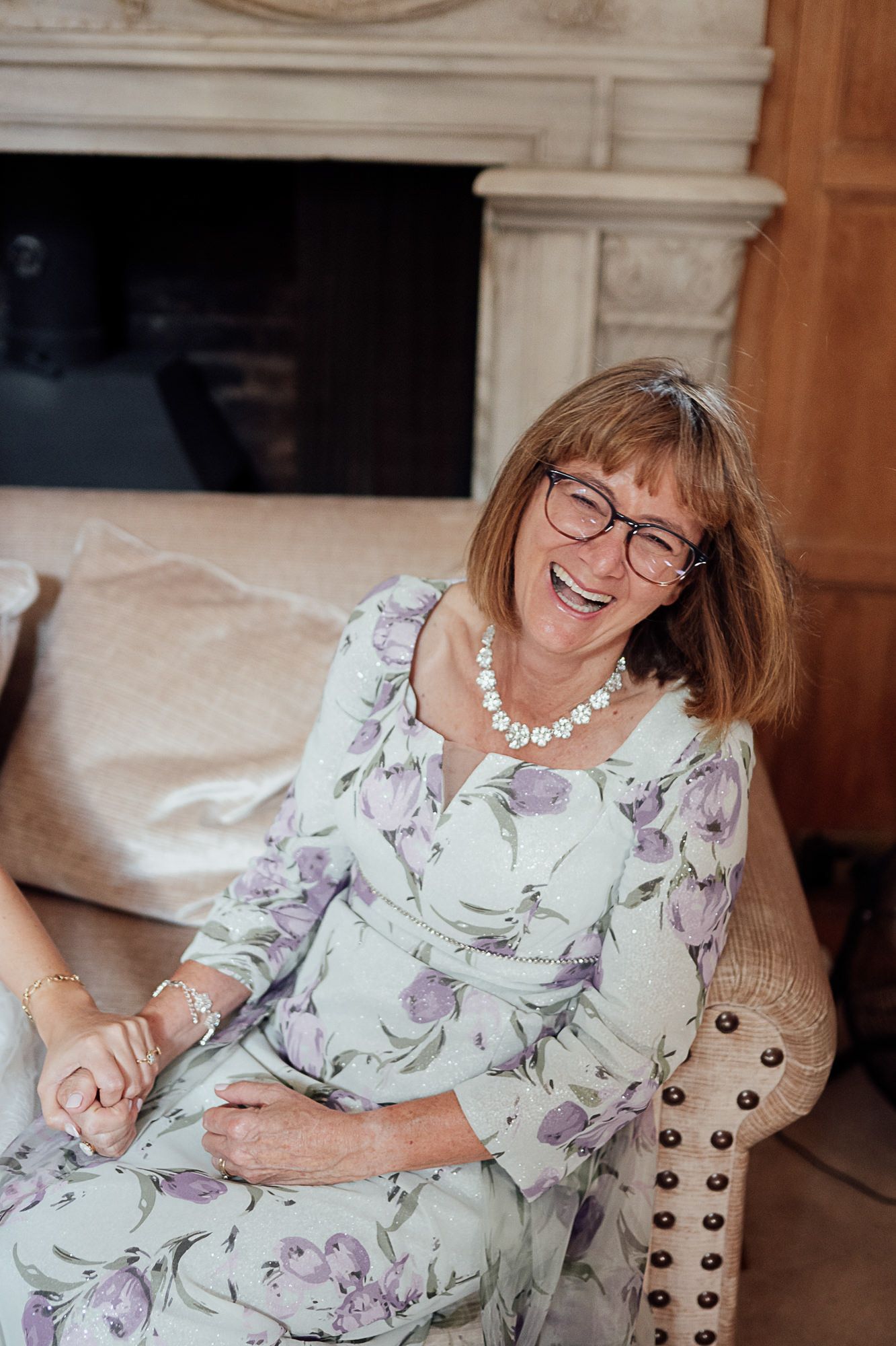 Smiling older woman in floral dress and pearl necklace laughing joyfully while seated on an elegant chair.