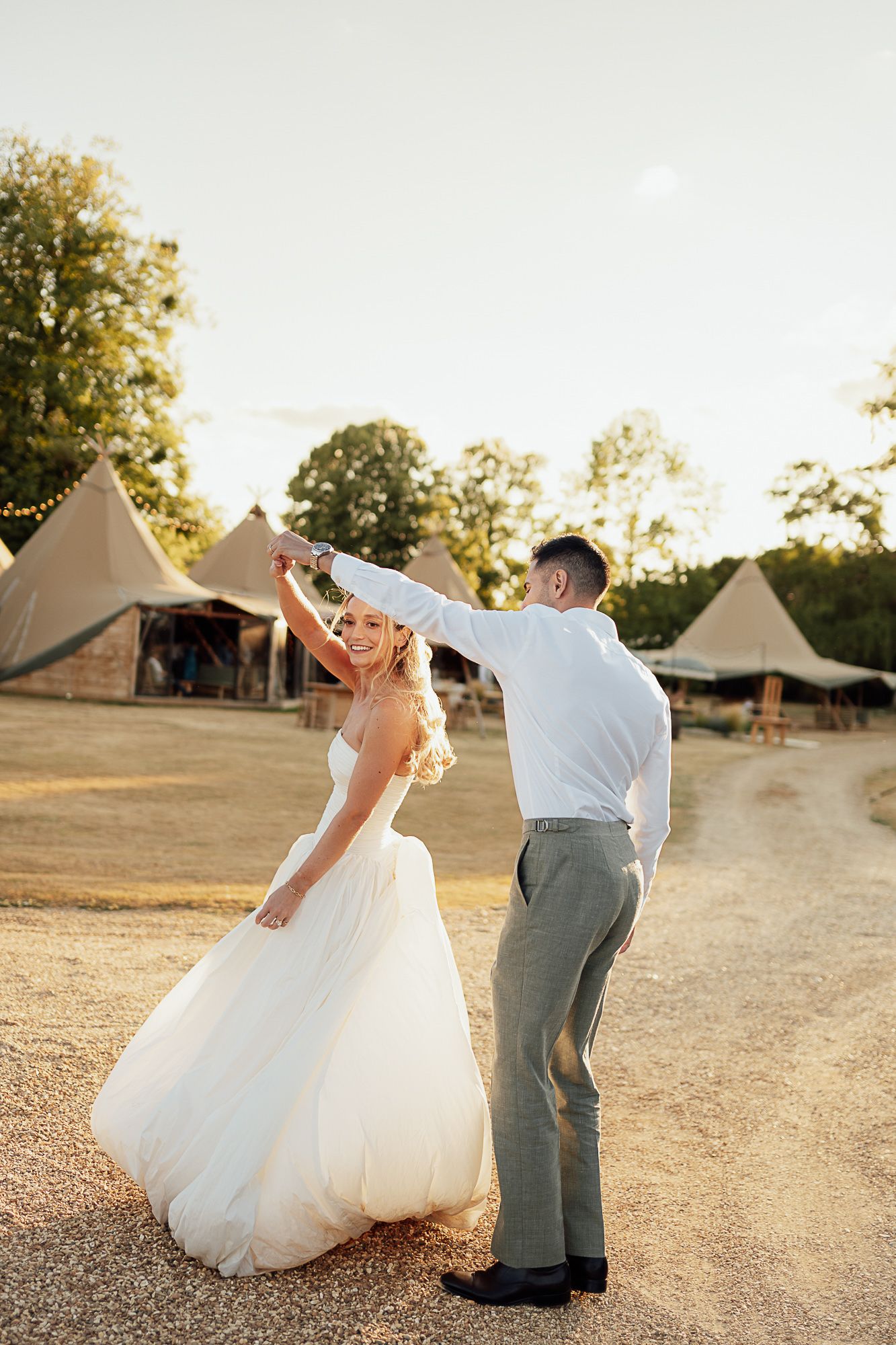 Bride and groom dancing outdoors at golden hour with tipi tents in background at rustic wedding venue.