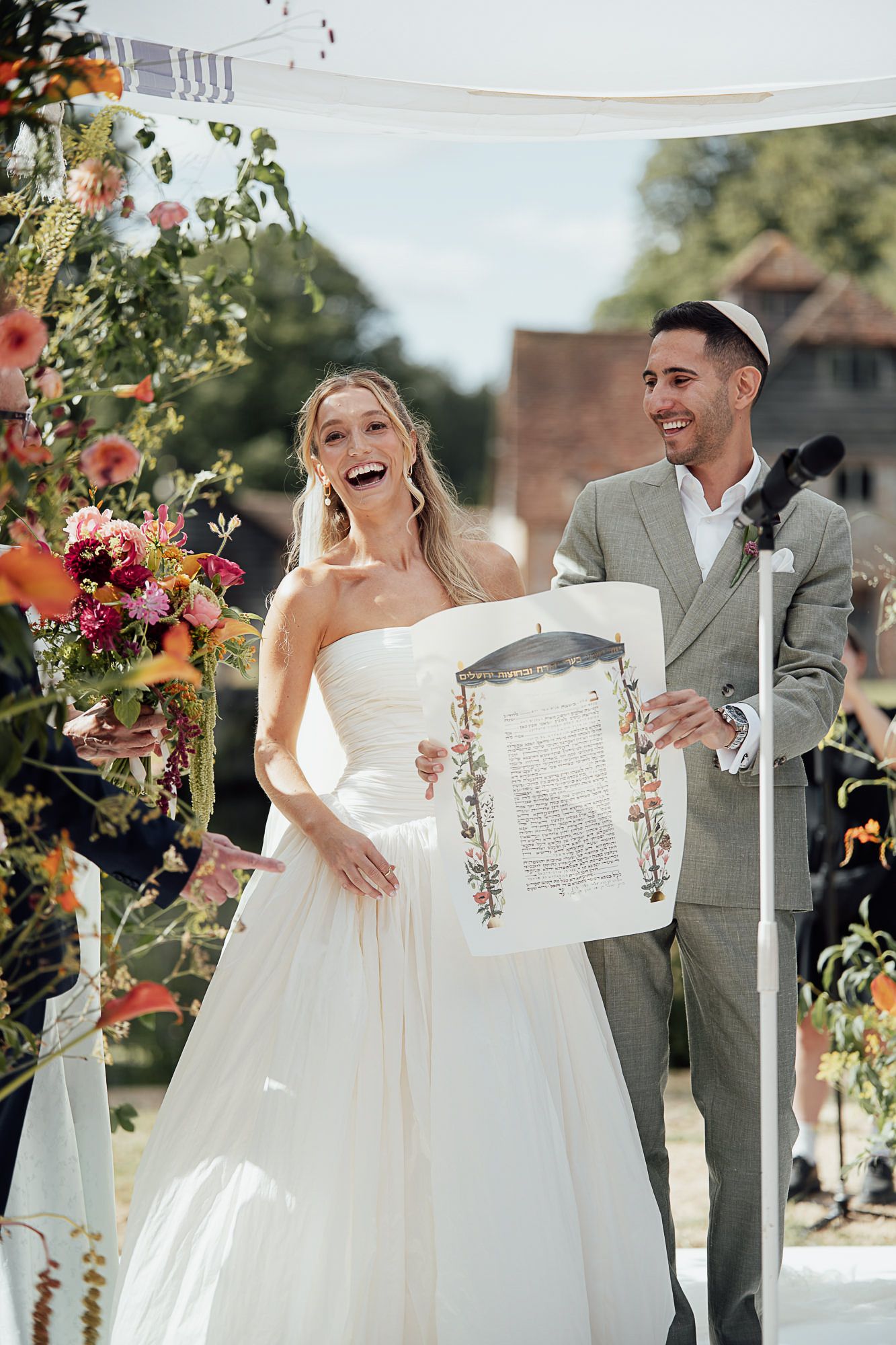 Joyful bride and groom hold ketubah under floral chuppah at outdoor Jewish wedding ceremony.