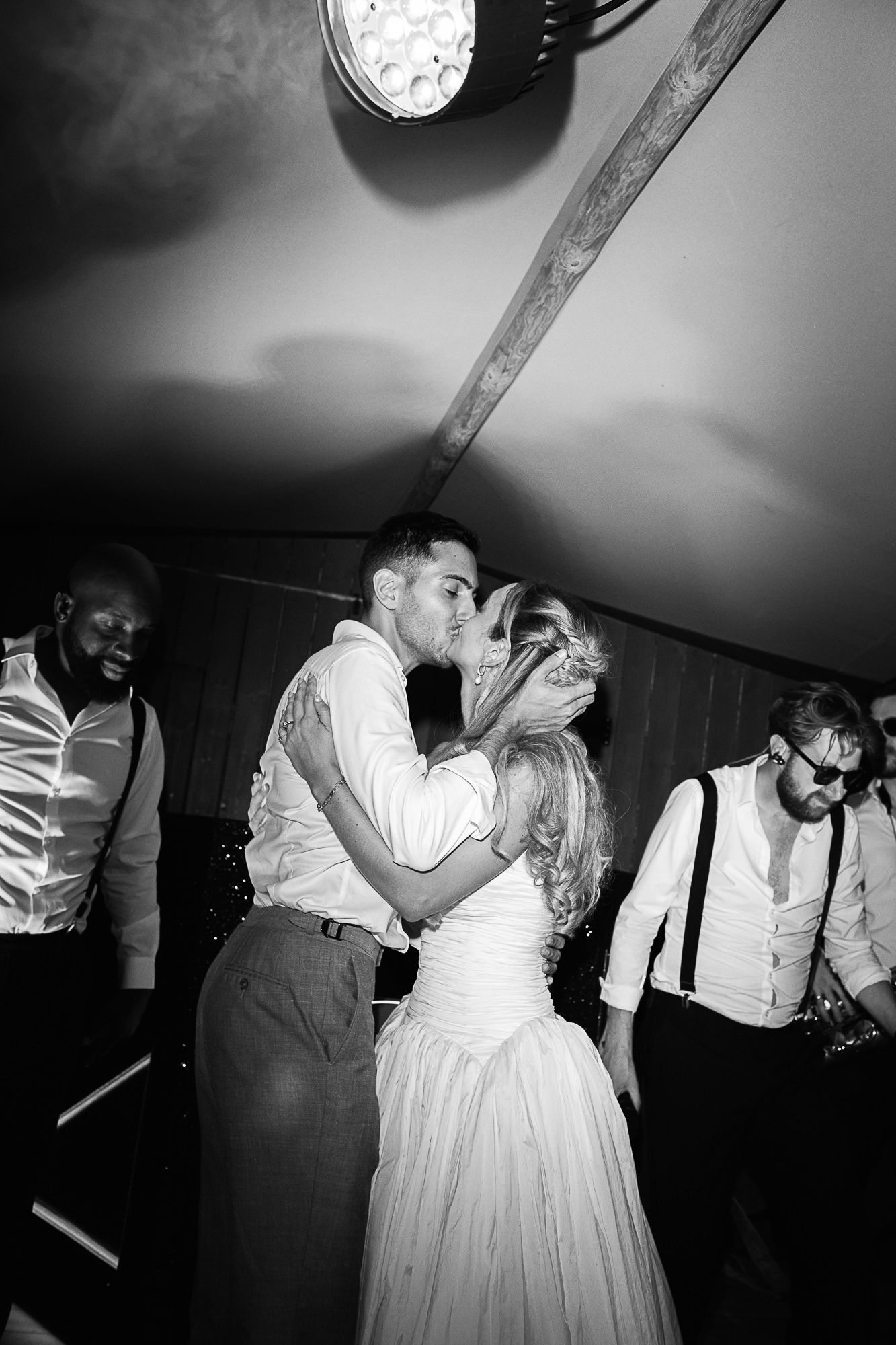 Black and white photo of a couple sharing a kiss on the wedding dance floor surrounded by guests.