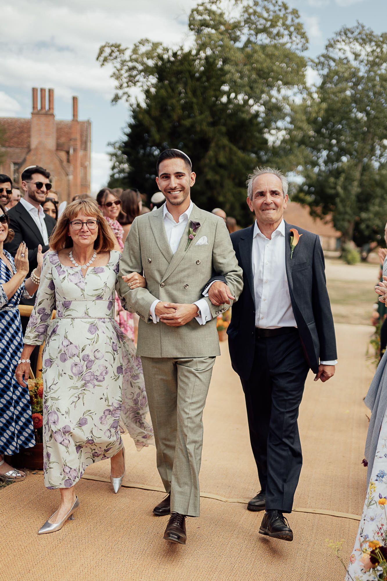 Groom in sage suit walks down aisle with parents at outdoor wedding ceremony near brick manor.