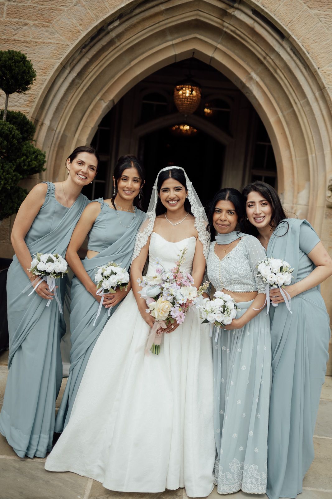 Bride in white gown with bridesmaids in dusty blue dresses holding bouquets outside a stone archway.