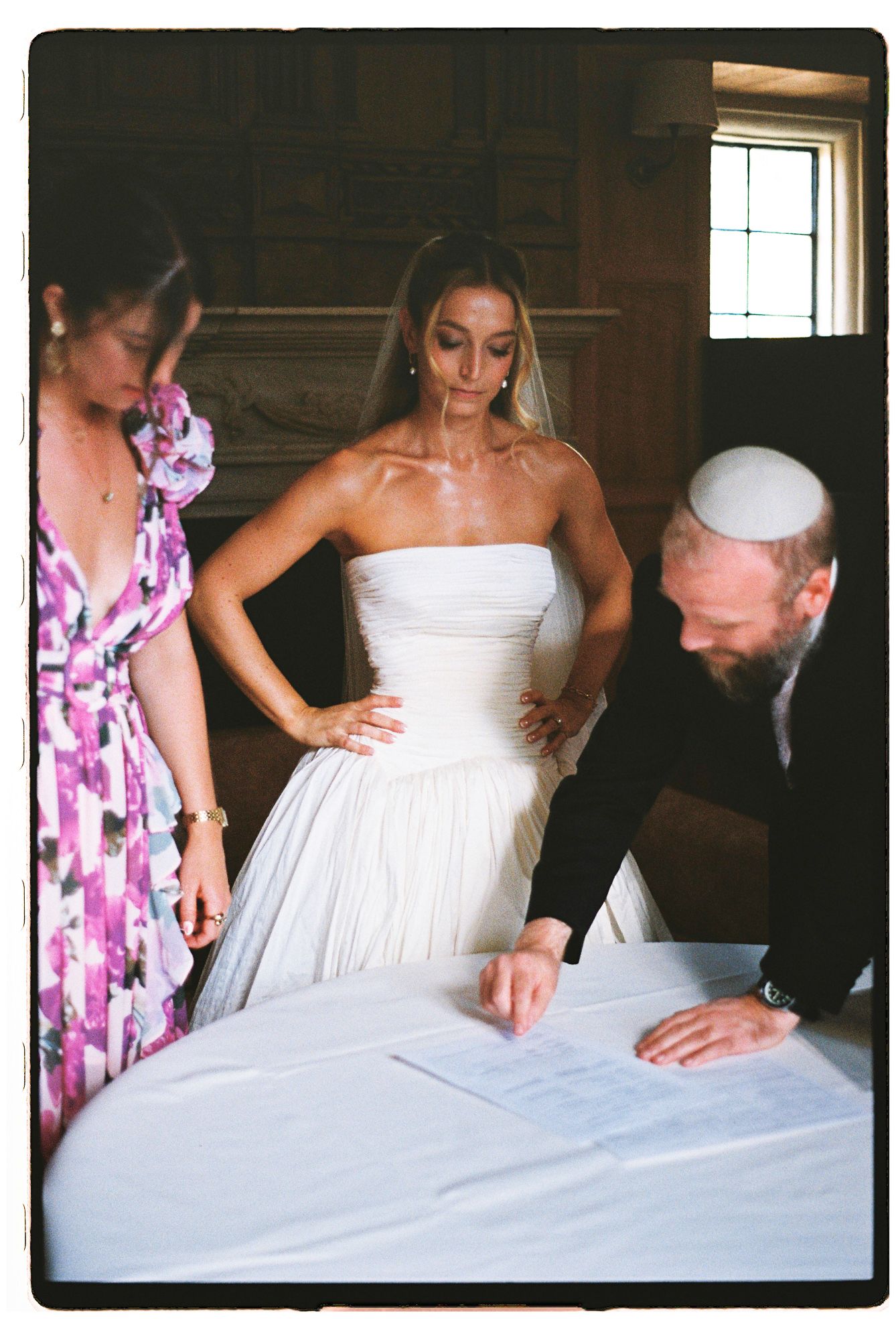 Bride in strapless white gown signing ketubah with rabbi in kippah during Jewish wedding ceremony.