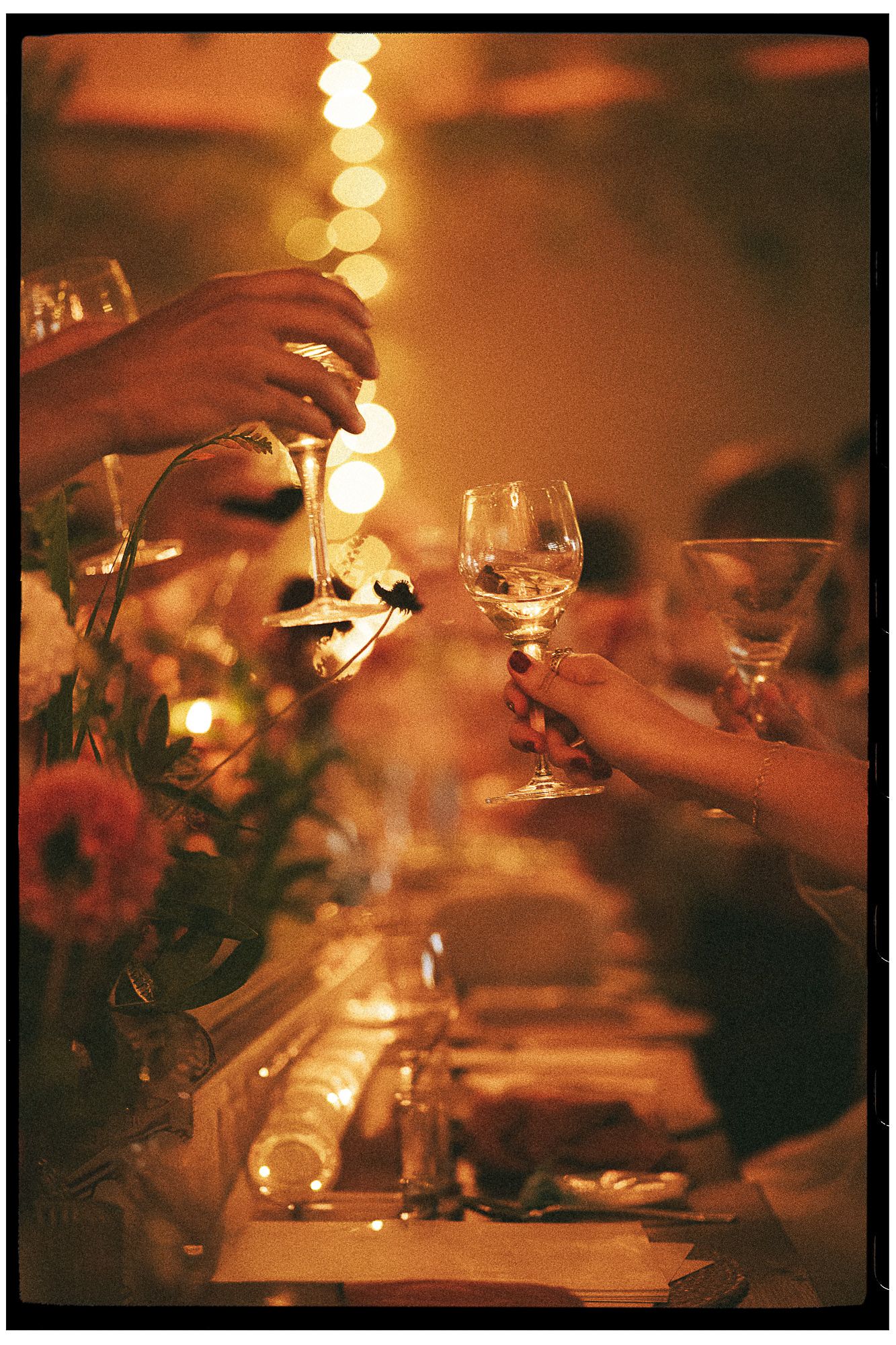 Elegant dinner party toast with wine glasses raised over candlelit table with floral centerpieces.