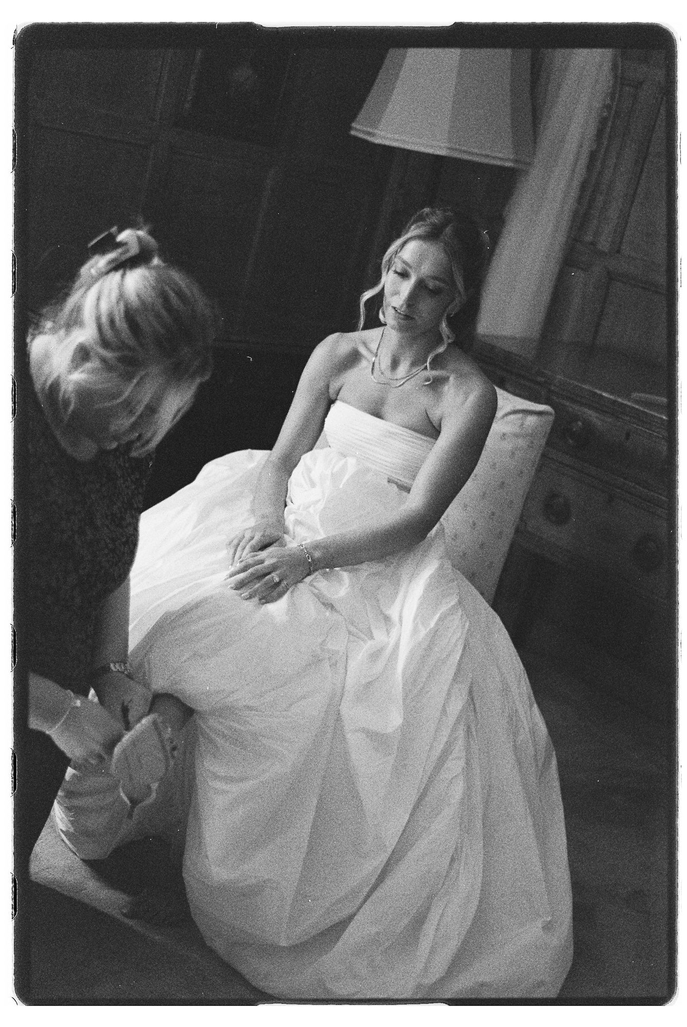 Black and white photo of a bride in a white gown seated while another woman assists her with her shoes.