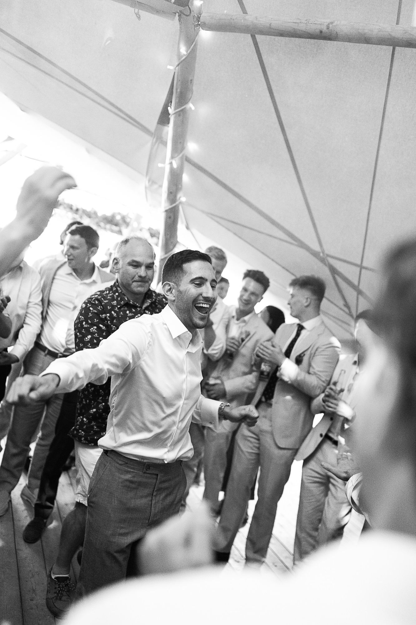 Black and white photo of men dancing joyfully at a wedding reception under a tent with string lights.
