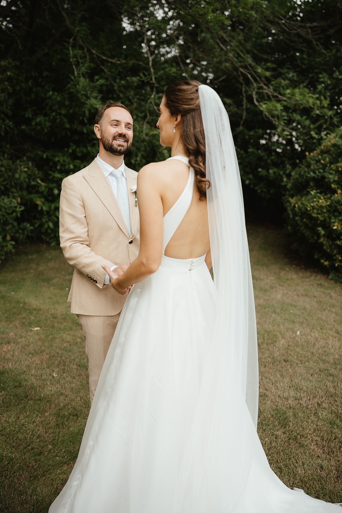 A joyful wedding couple stands together outdoors, with the bride in a backless white gown and flowing veil.