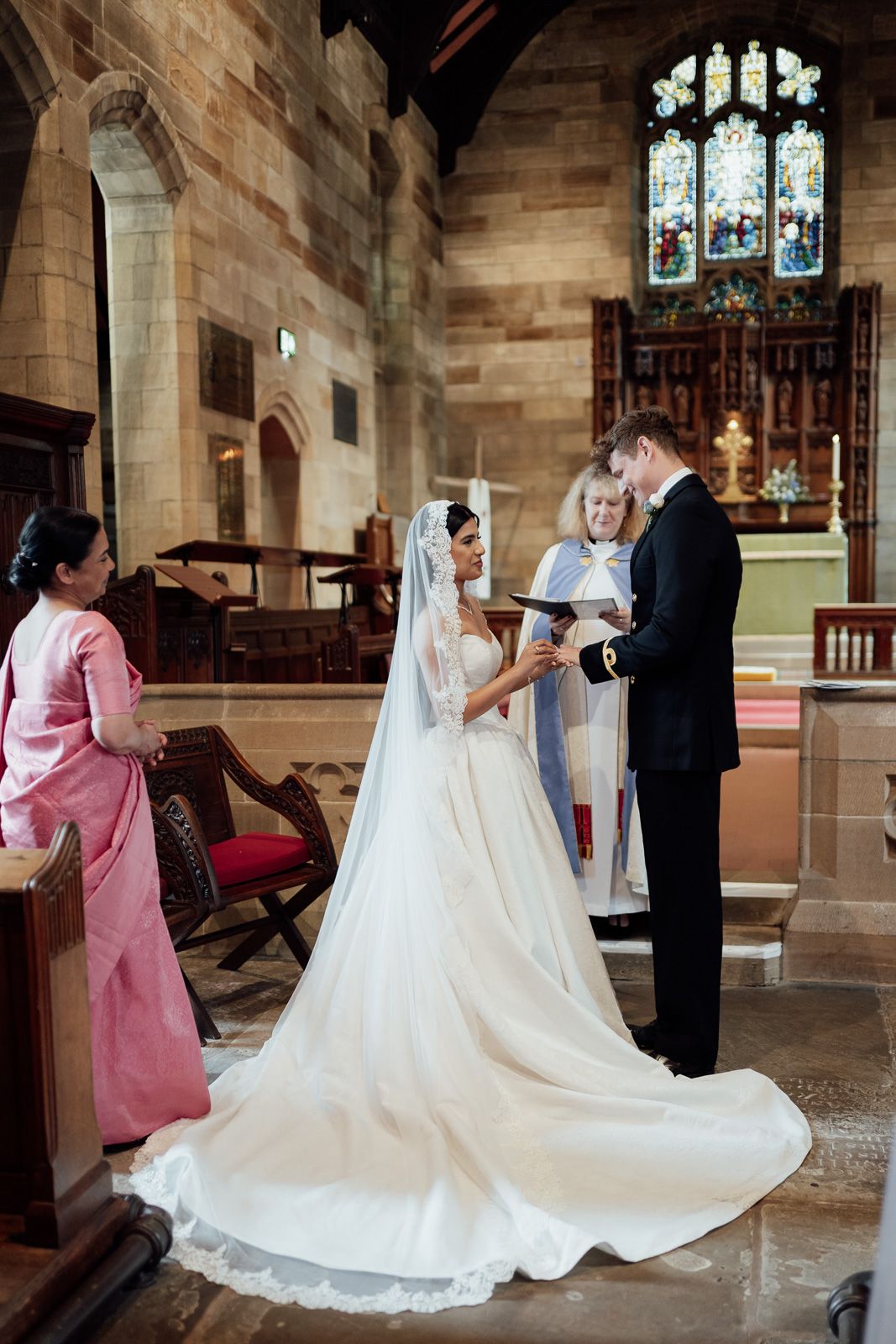 Bride and groom exchange vows at a church altar, officiated by a minister, with a guest in pink watching nearby.