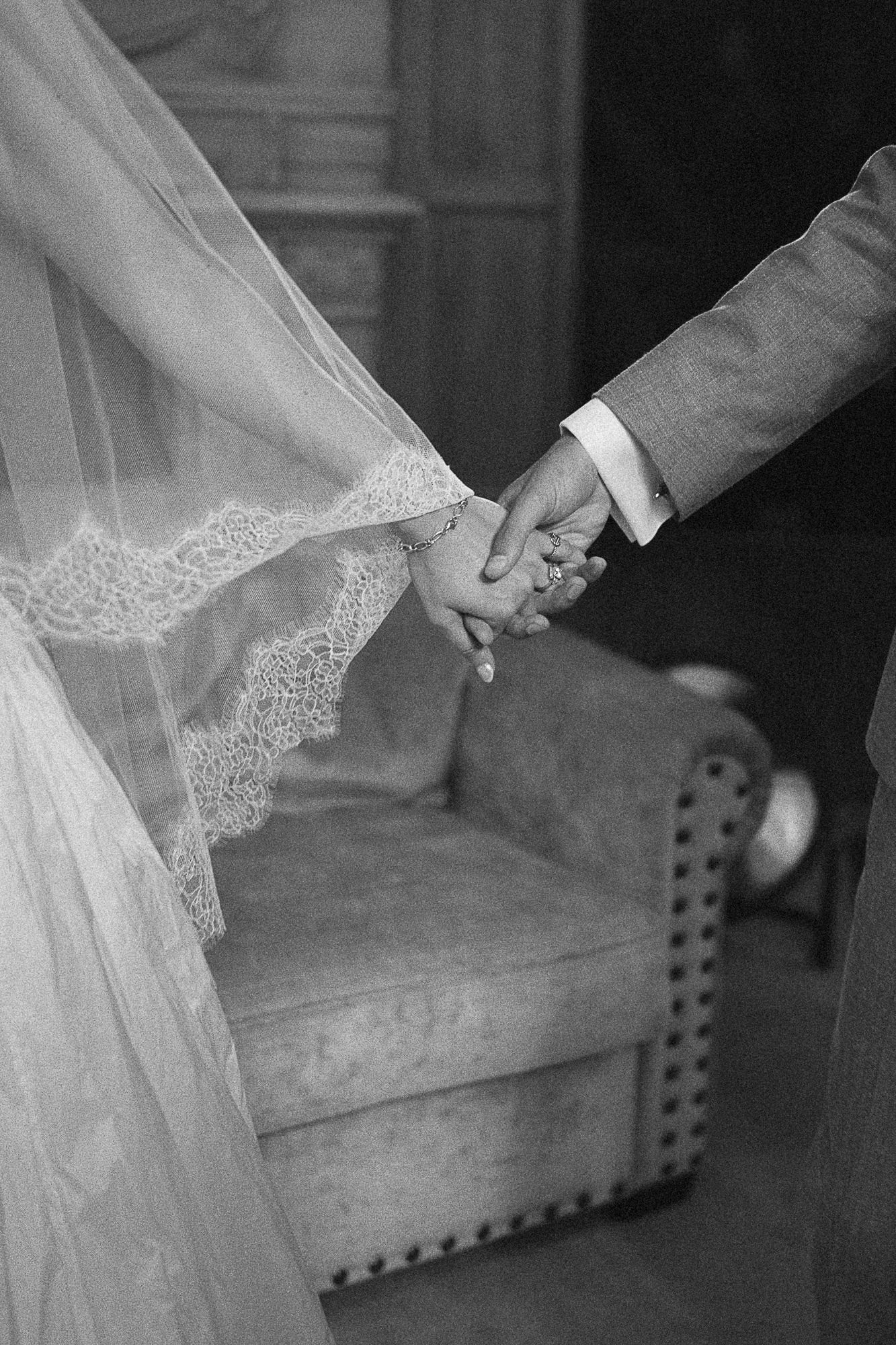 Black and white photo of bride and groom holding hands, wedding veil and suit visible, elegant chair in background.