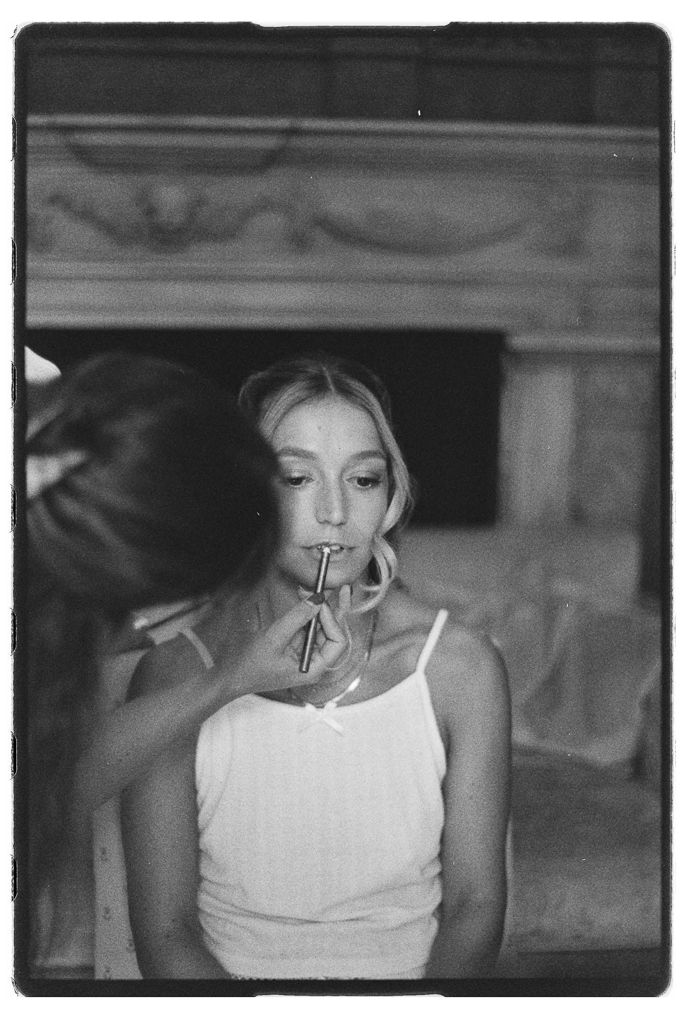 Black and white film photo of a young woman in a white tank top having her makeup applied by an artist.