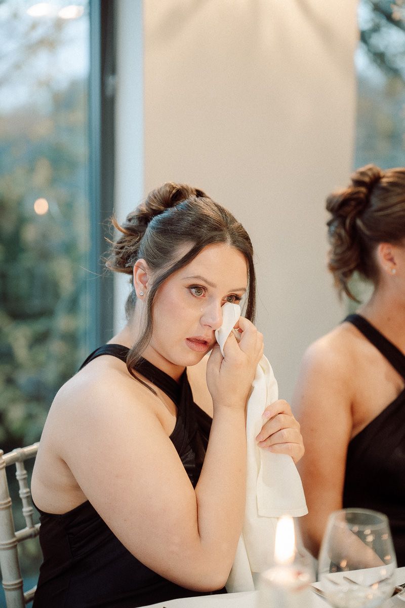 A person in a black dress wipes tears away with a napkin while seated at a formal event.