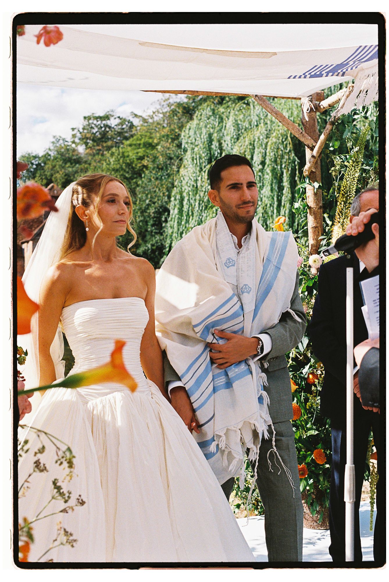 Bride and groom stand under a chuppah during an outdoor Jewish wedding ceremony.