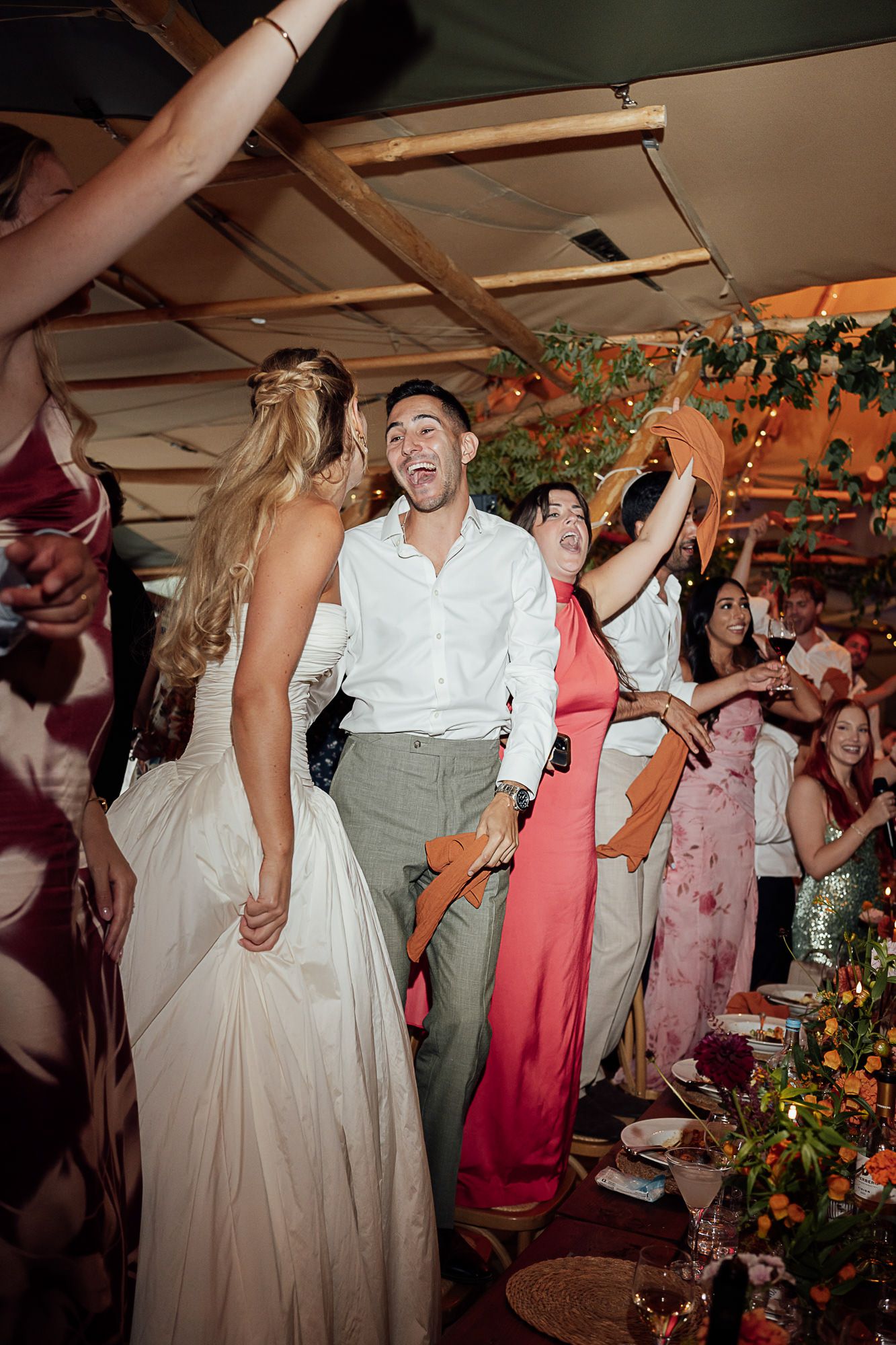 Bride and groom laughing and dancing joyfully with guests at a lively indoor wedding reception celebration.