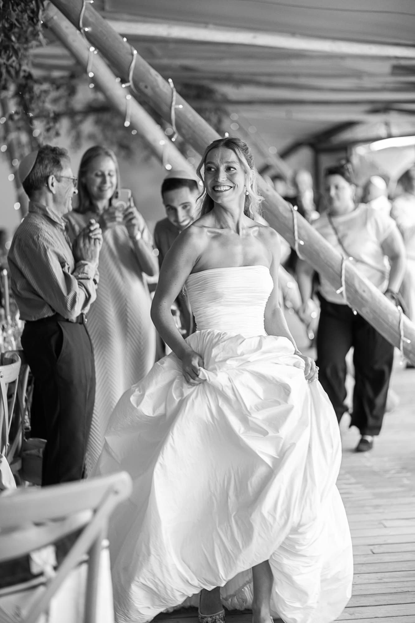 Black and white photo of a radiant bride in a strapless gown walking joyfully at her wedding reception.