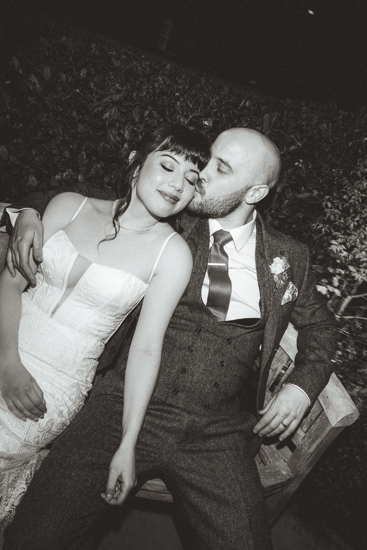 Black and white photo of a romantic wedding moment with couple embracing outdoors, bride in lace dress and groom in suit.