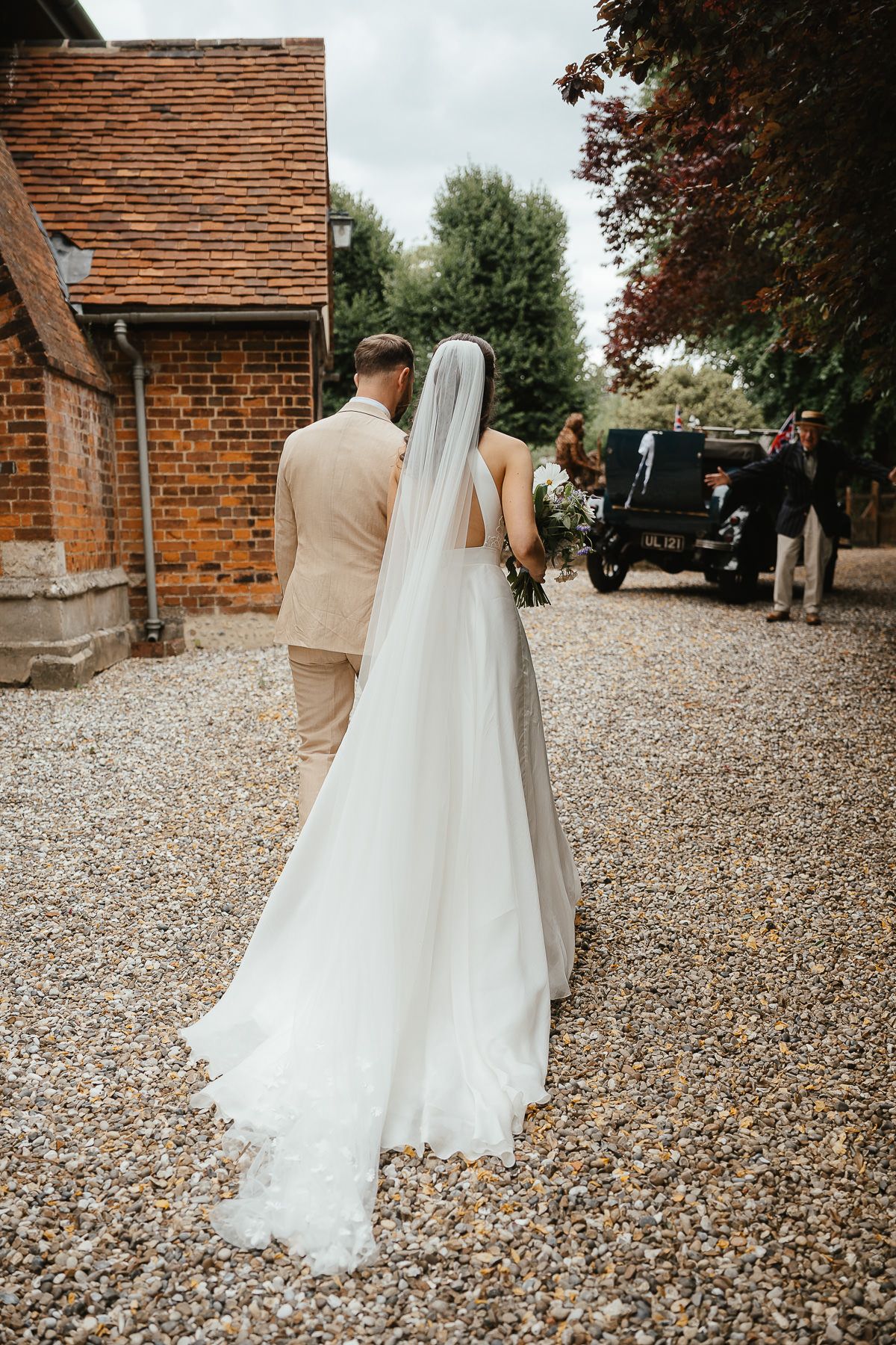 A bride in a flowing white veil and groom walk on gravel path towards vintage black car at rustic brick venue.