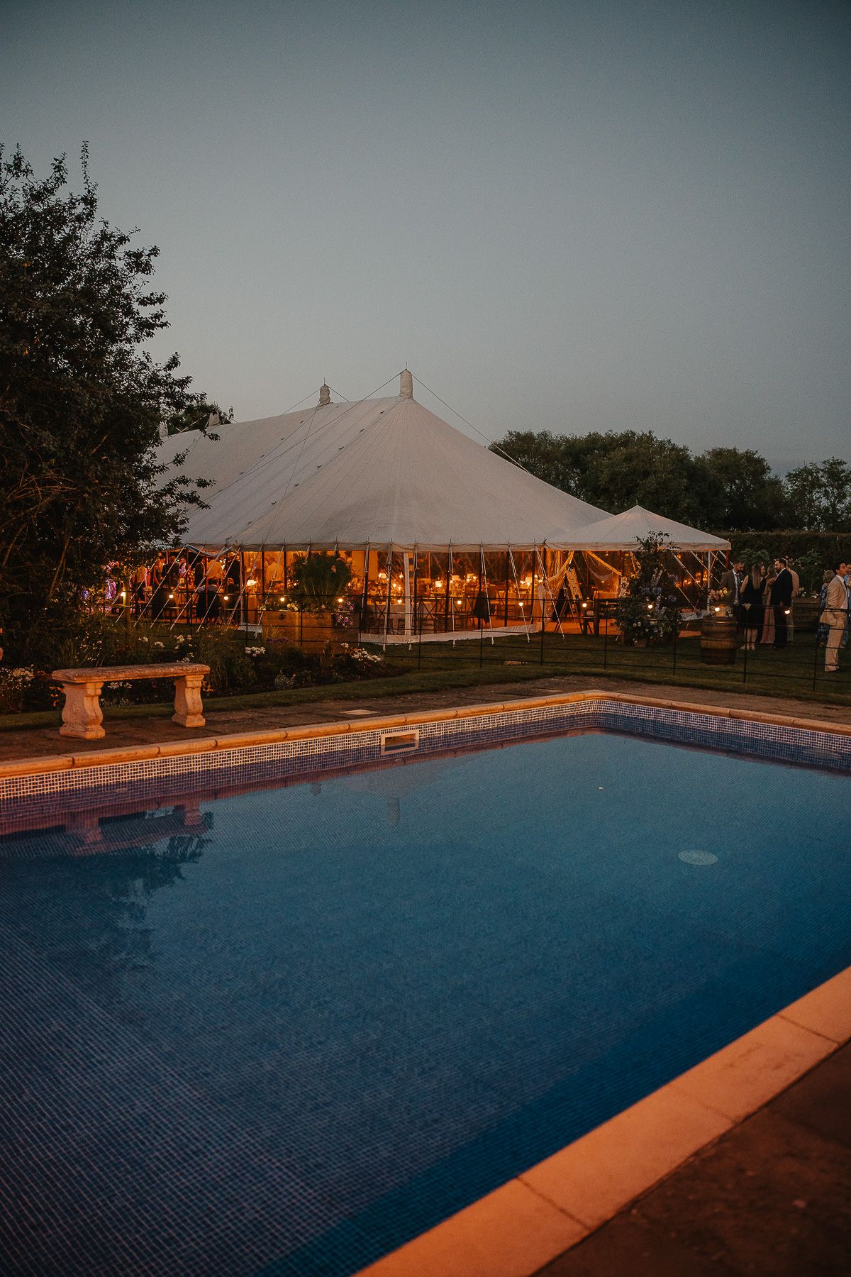 Illuminated wedding tent glows at dusk beside a swimming pool during an elegant outdoor evening reception.