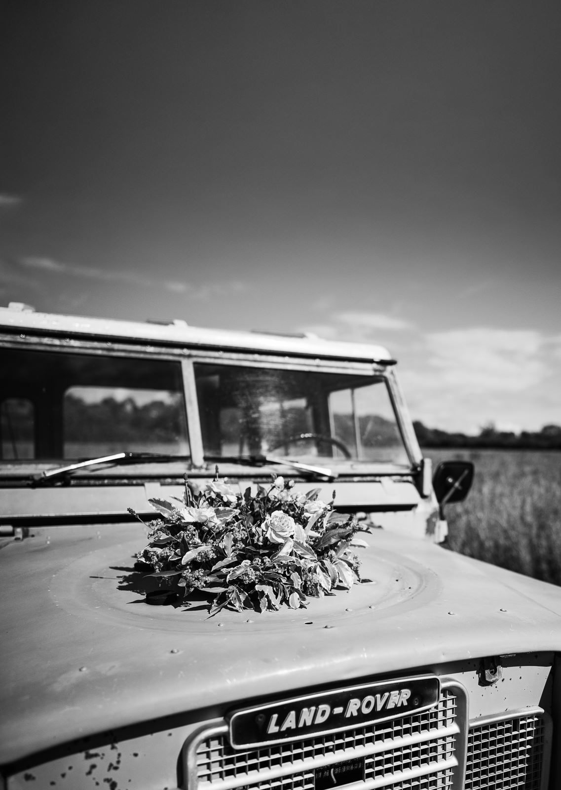 A black and white photo of a classic Land Rover with a floral bouquet displayed on its hood in a rural setting.