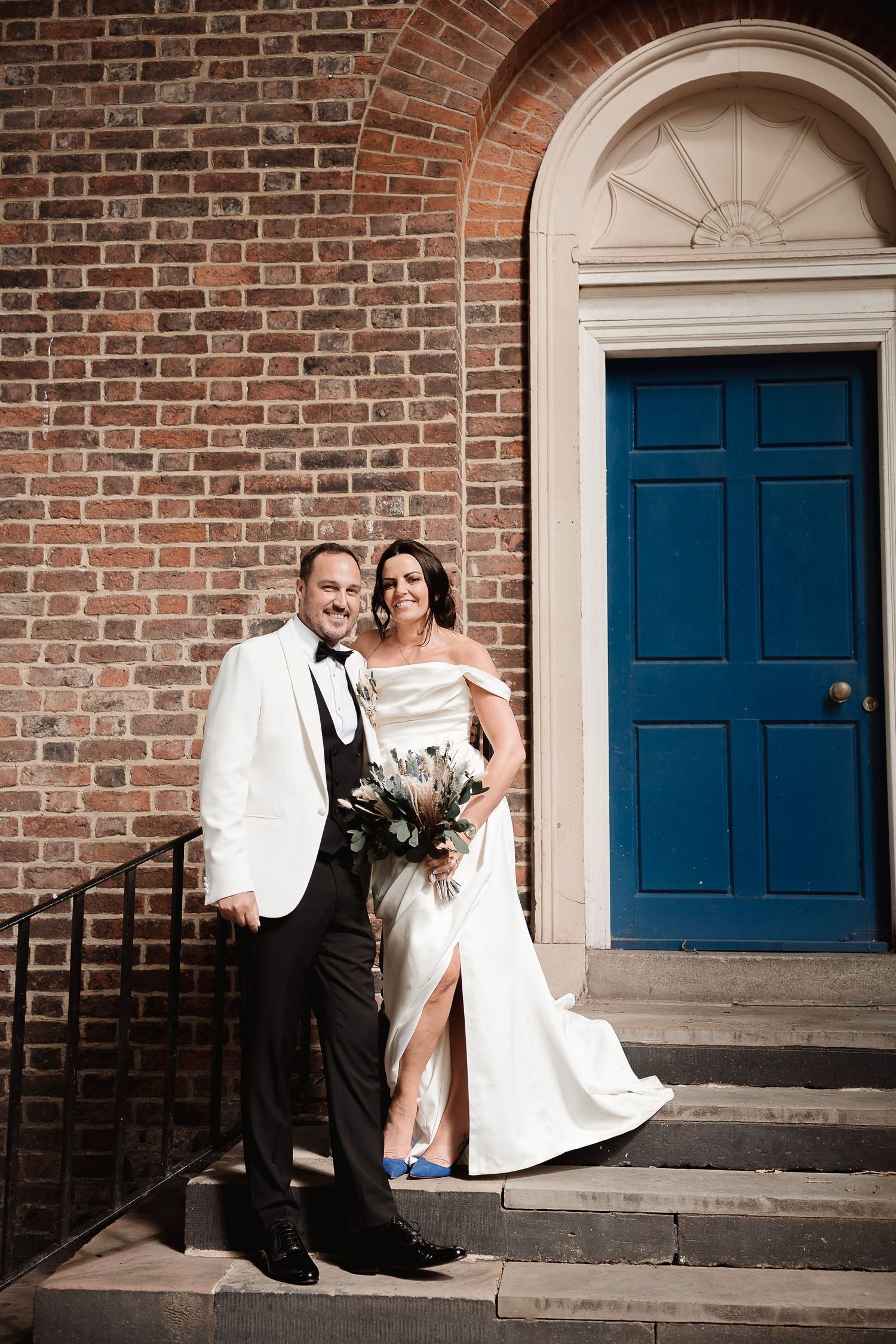 A couple poses in formal wedding attire in front of a historic brick building with a bright blue door.