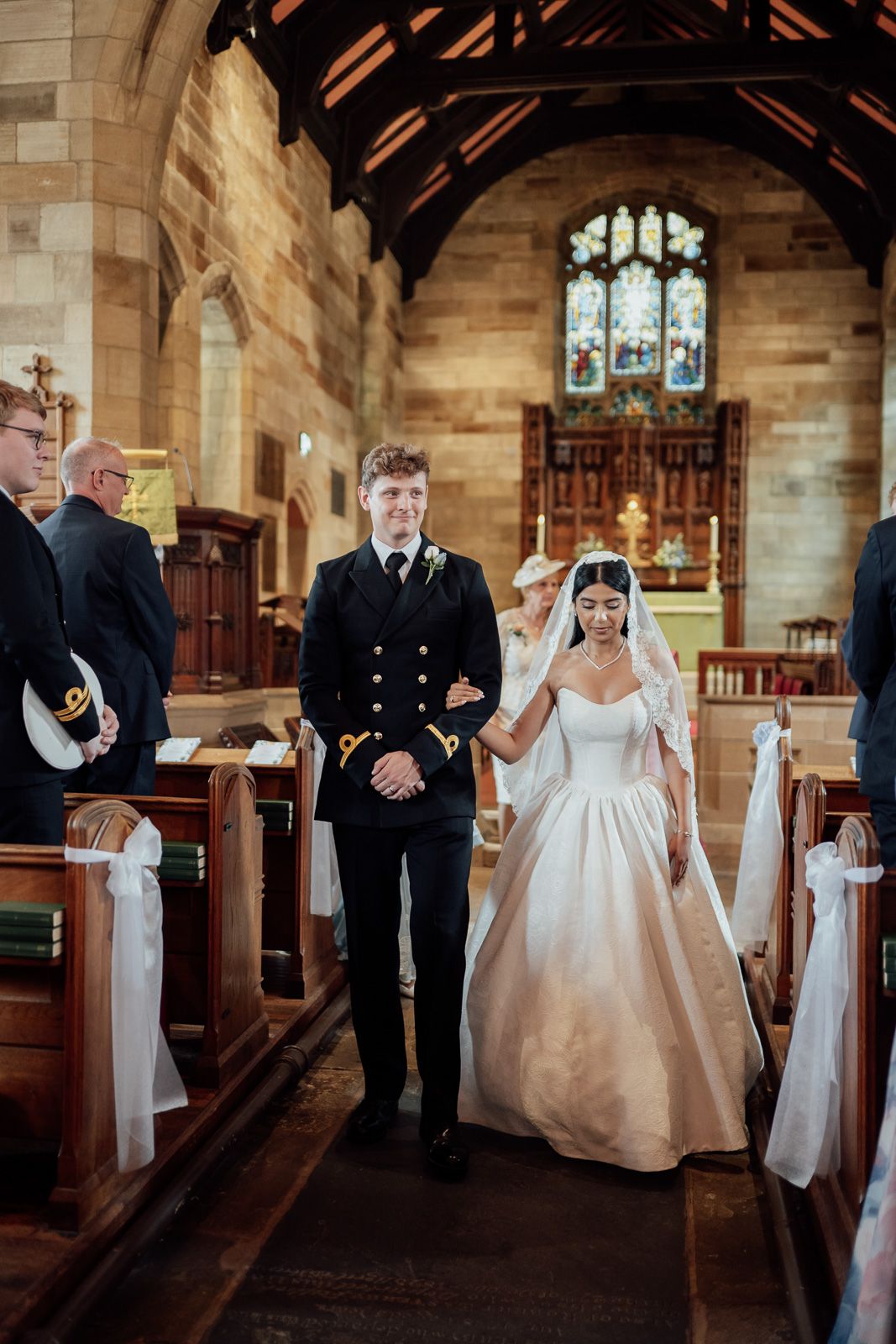 Bride and groom walk down the aisle of a historic stone church, groom in naval uniform.
