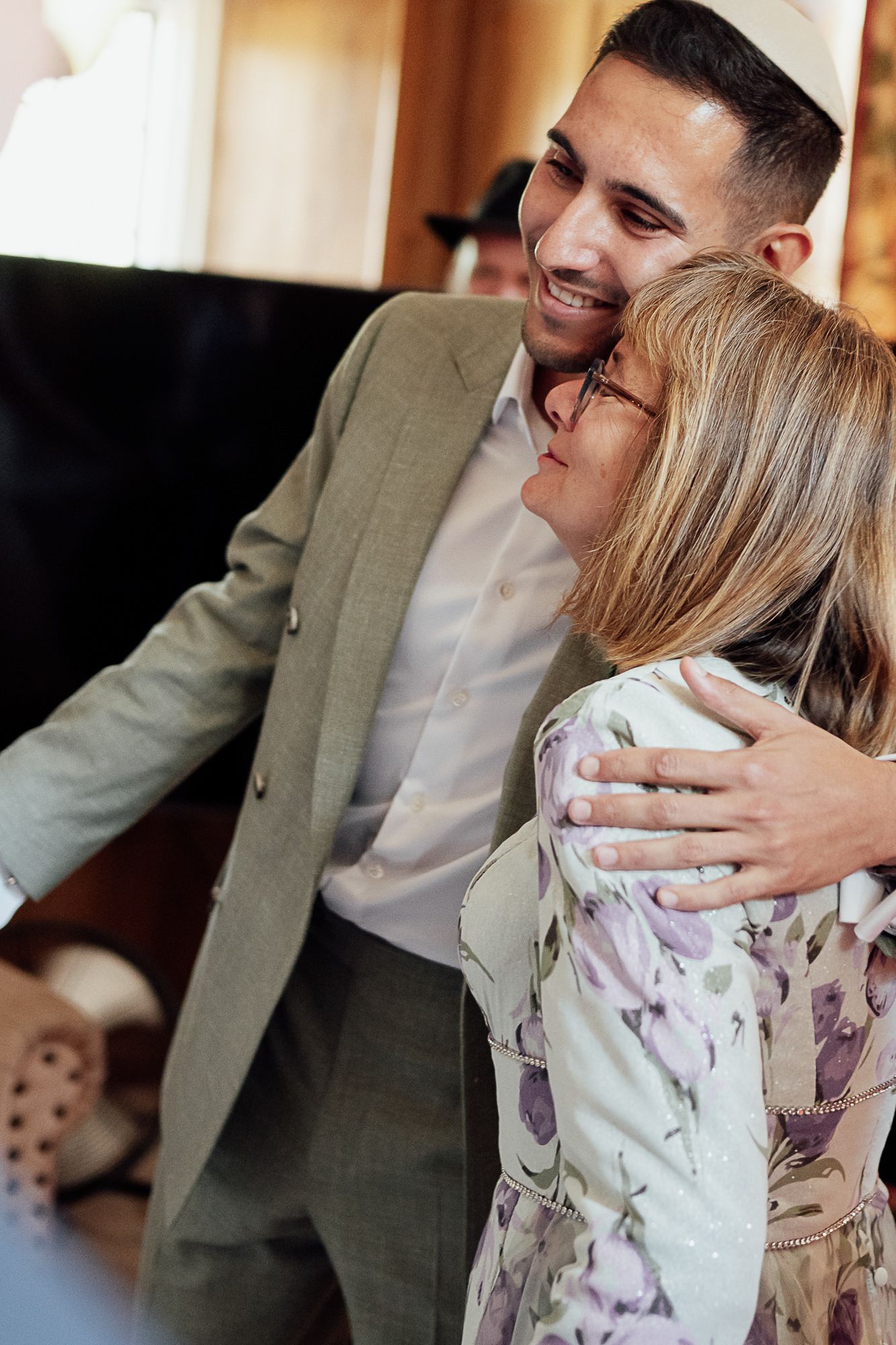 A smiling young man in a gray suit embraces an older woman with glasses in a floral blouse indoors.