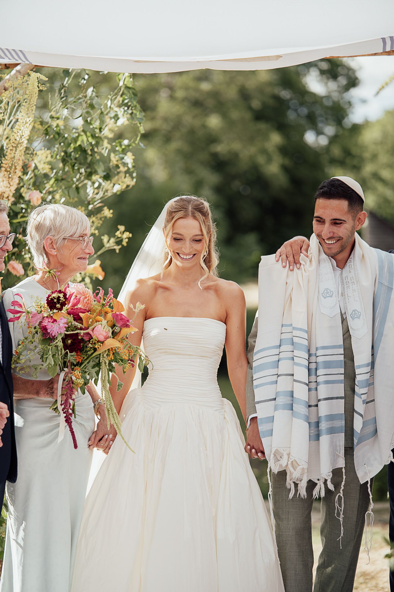 Smiling bride in white gown holds hands with groom wearing tallit under outdoor wedding chuppah.