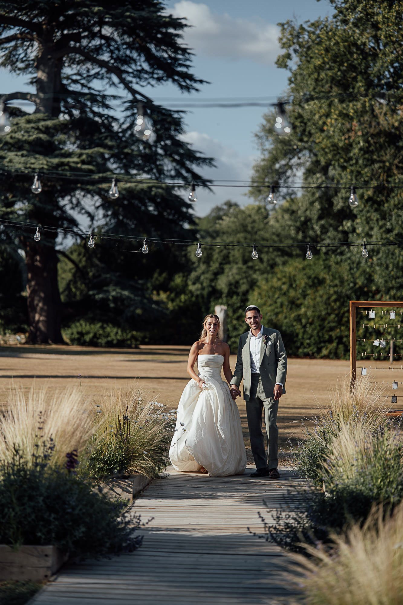 Bride and groom walk hand in hand down a wooden path surrounded by ornamental grasses and string lights at an outdoor wedding.