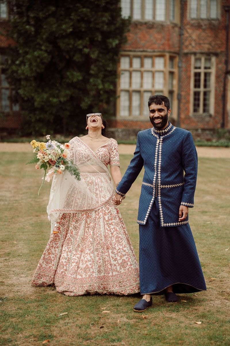 A couple in traditional wedding attire, with a pink embroidered gown and navy military-style jacket, pose in a garden setting.