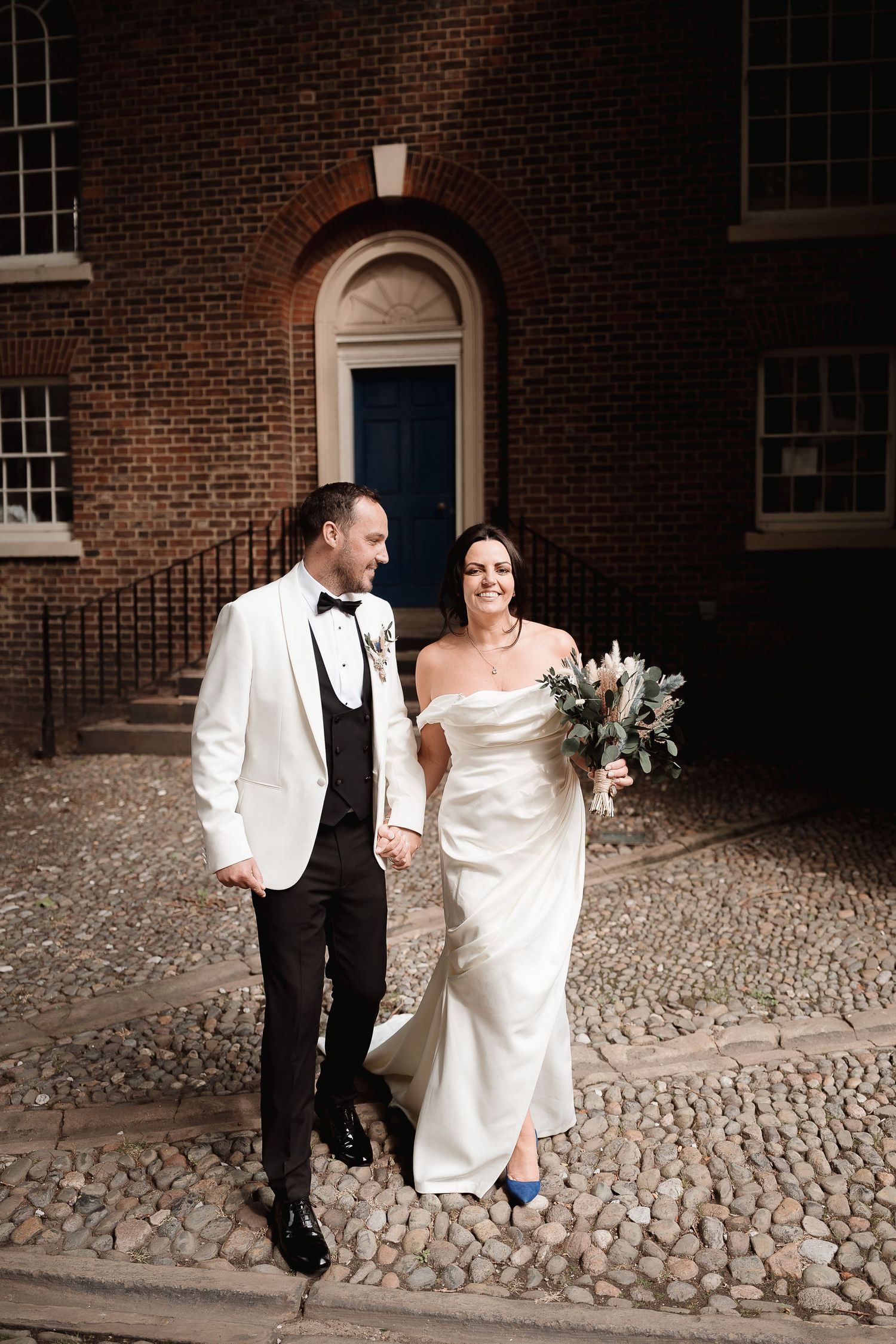 A couple in wedding attire stands in front of a historic brick building with an arched doorway on a cobblestone path.