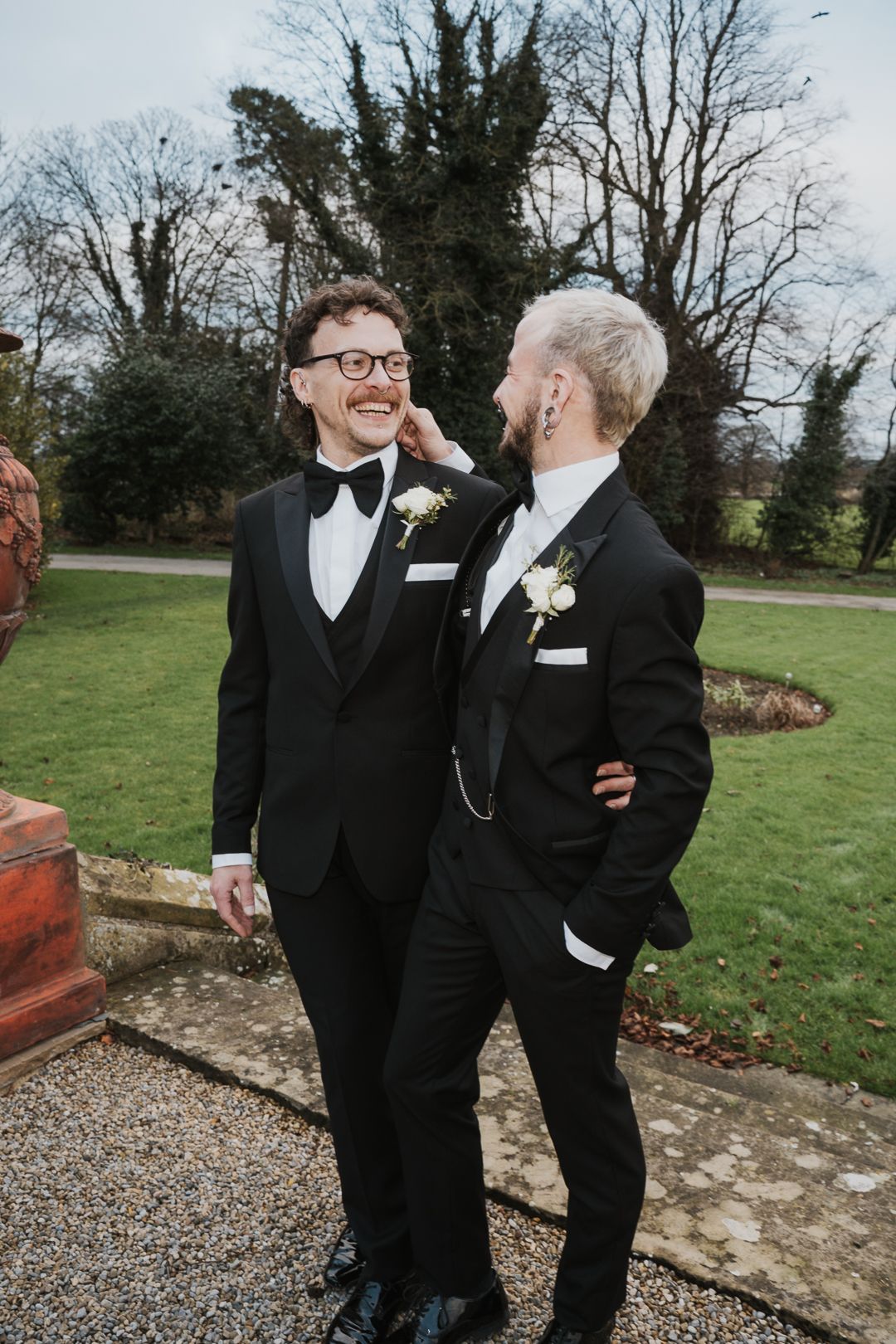 Two wedding guests in black tuxedos and bowties share a joyful moment together on a lawn with bare trees in background.