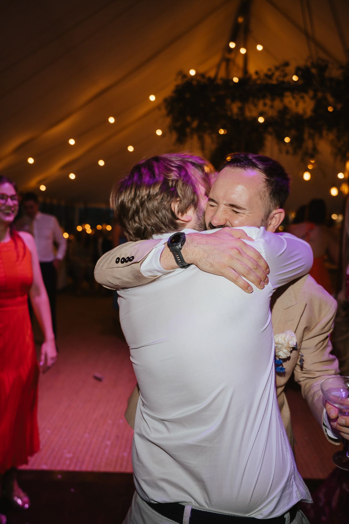 Two guests embrace emotionally on a dance floor under warm string lights at an evening wedding reception.