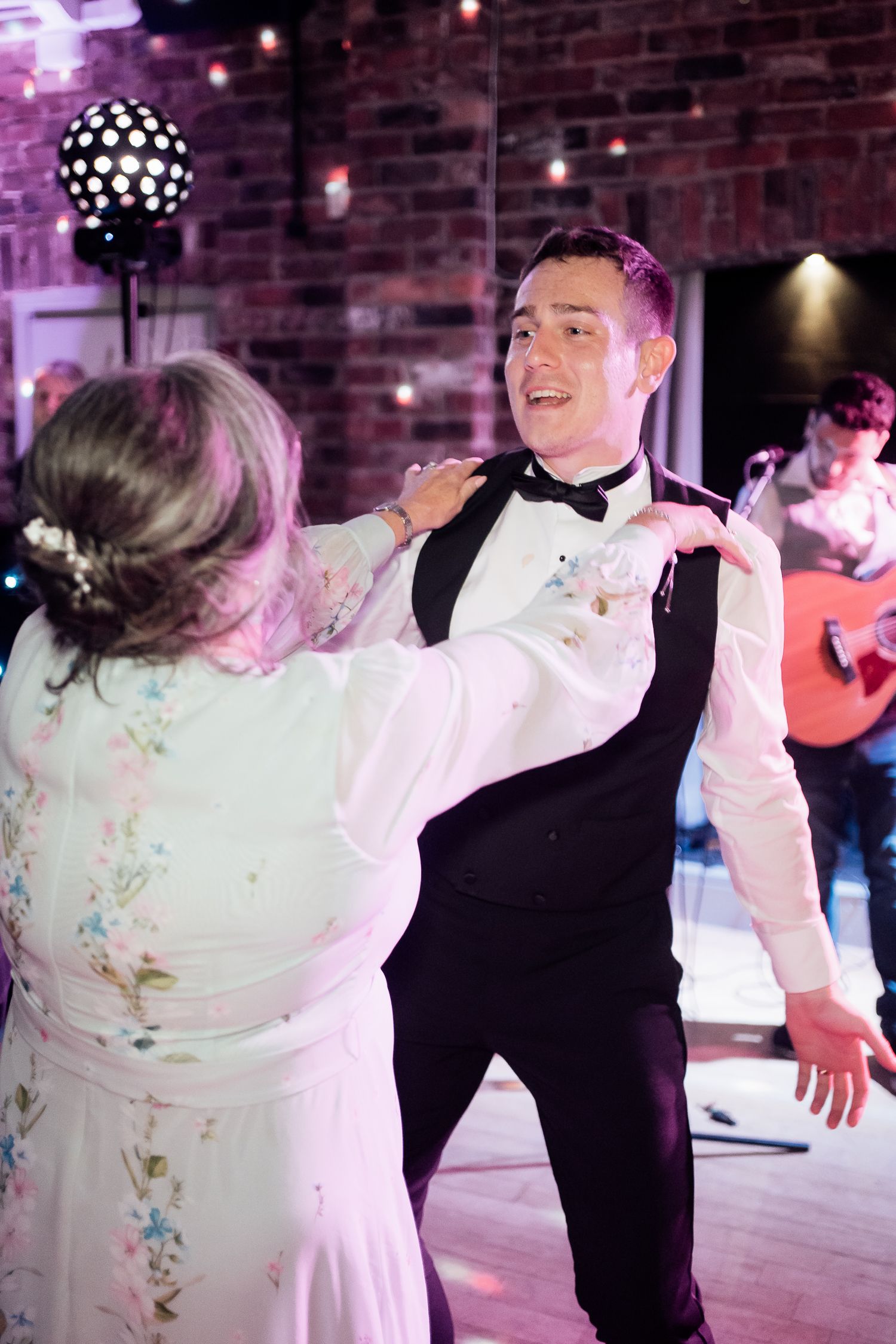 A couple dances under a disco ball at what appears to be a wedding reception with a rustic brick wall backdrop.