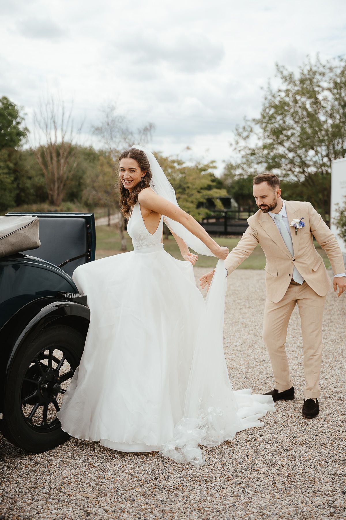 Couple poses by vintage black car on their wedding day with bride in white gown and groom in tan suit.