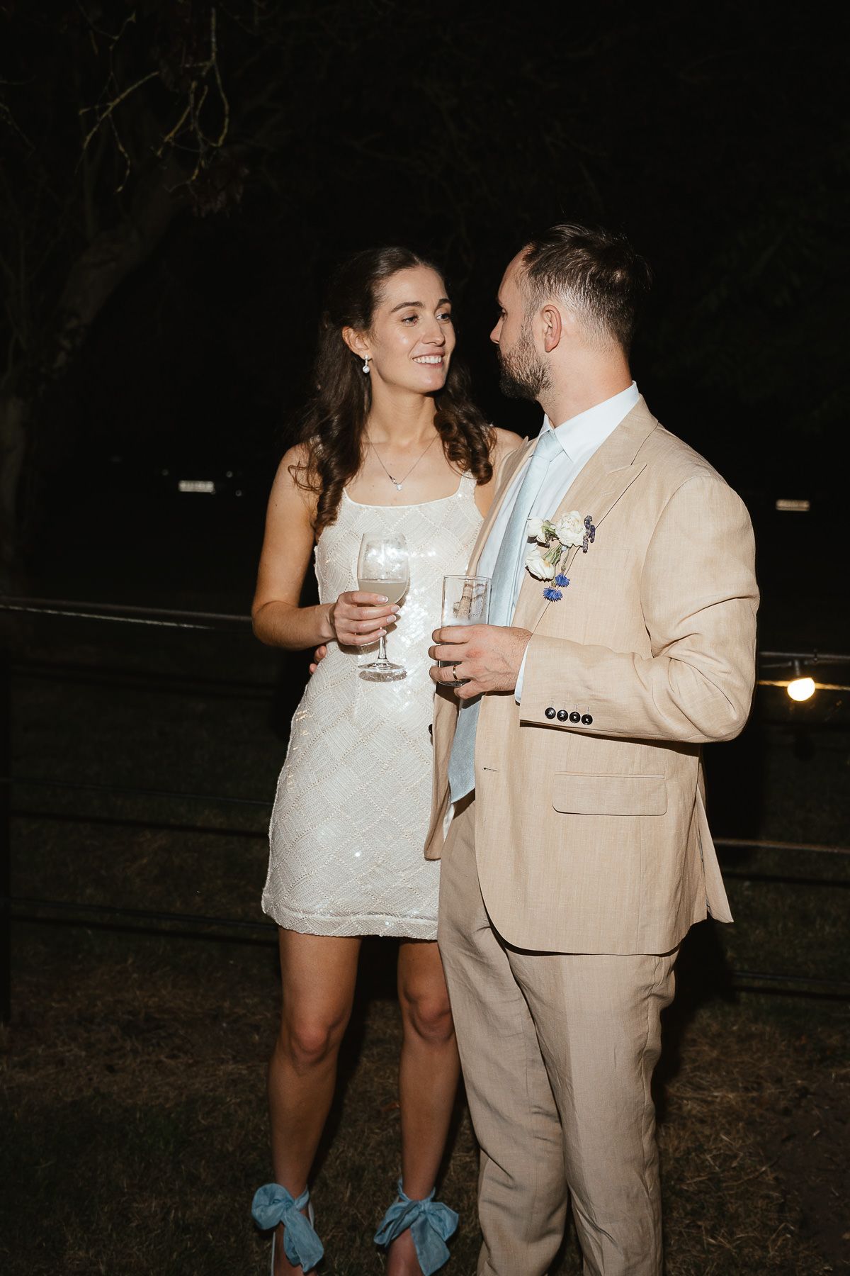 A couple in wedding attire sharing drinks and intimate moments at a nighttime outdoor reception.