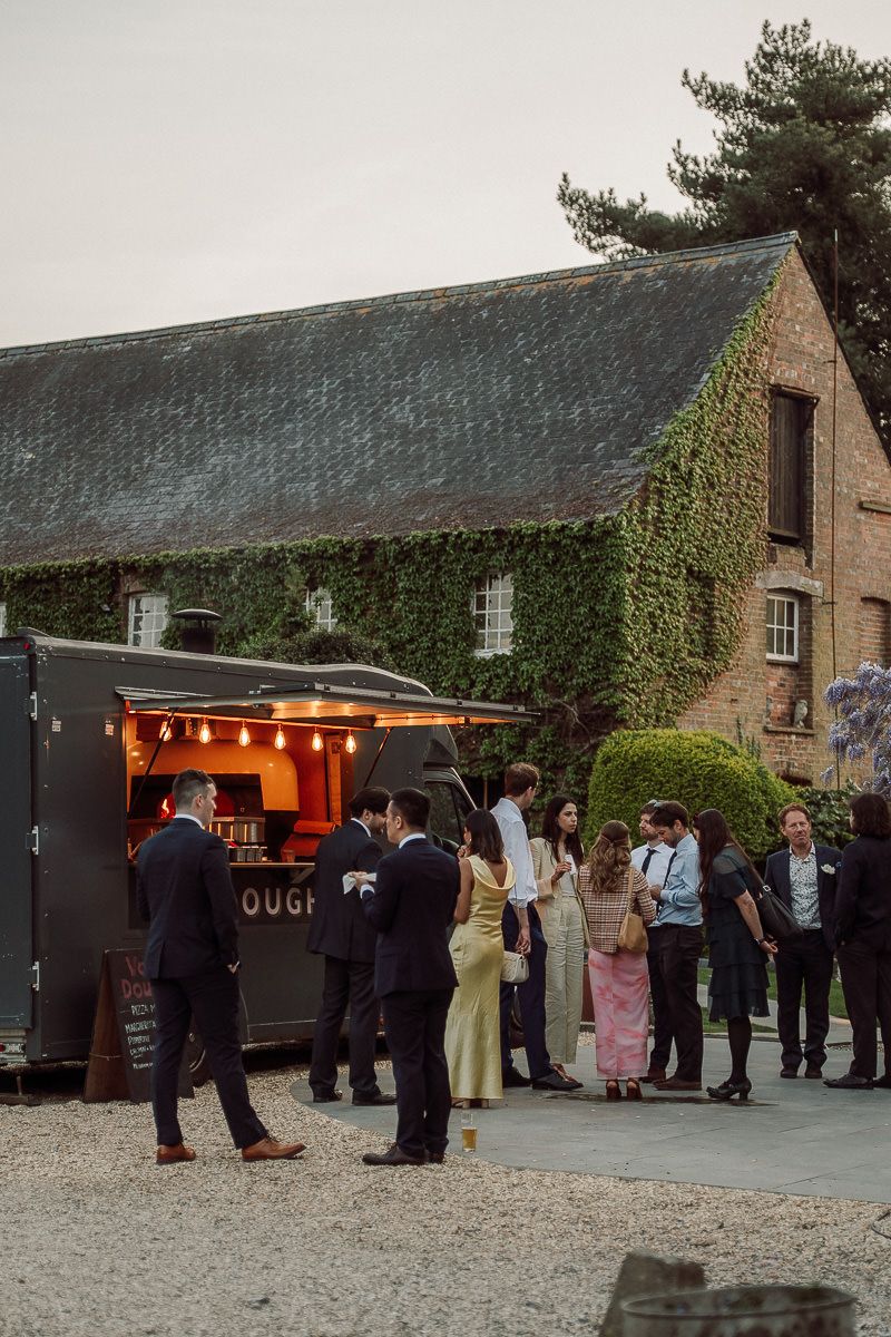 People gather at a rustic stone building with an illuminated food truck serving refreshments at dusk.