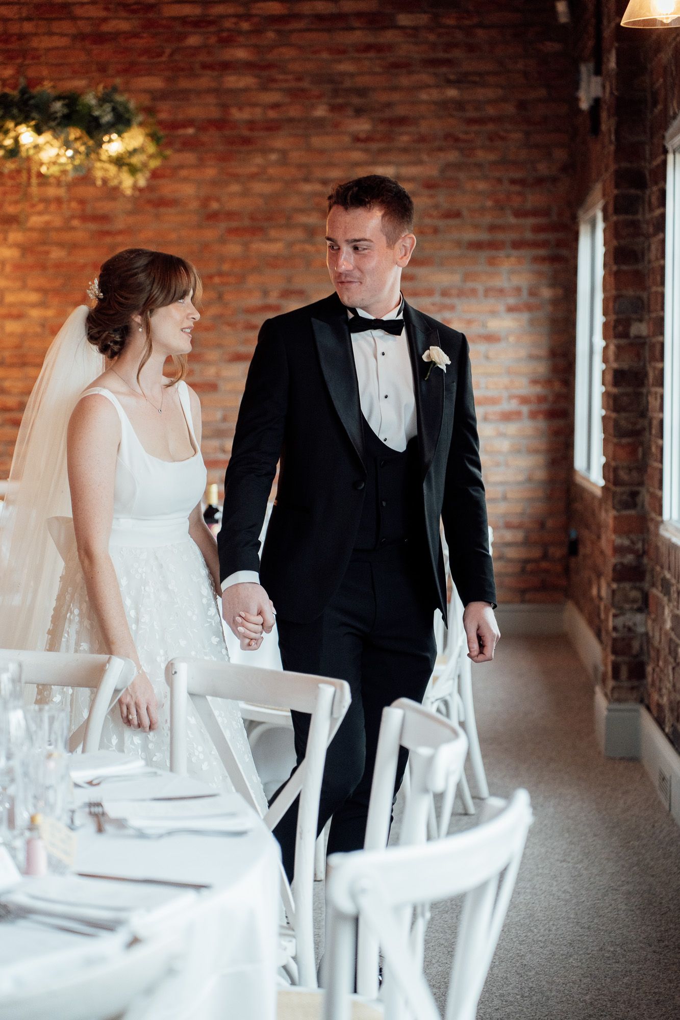 A bride and groom holding hands by an elegantly set dining table with a brick wall backdrop and white chairs.