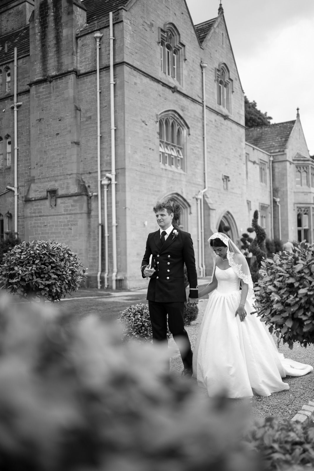 Black and white photo of a bride and groom walking outside a historic stone building on their wedding day.