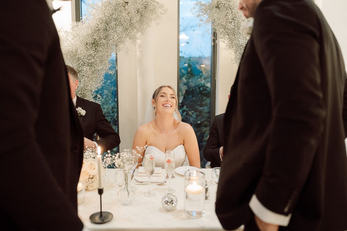 A bride seated at a wedding reception table laughs while guests in dark suits stand nearby.
