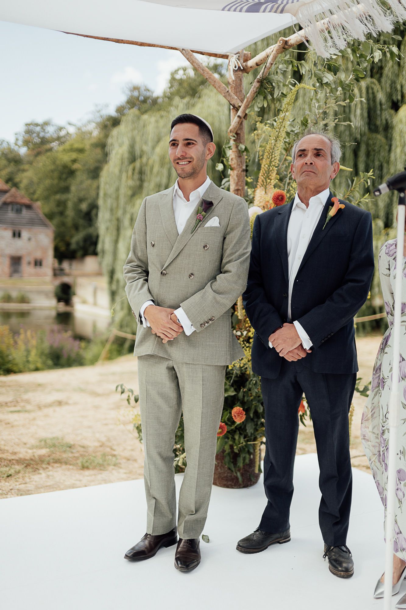 Groom in light gray double-breasted suit stands with older man in navy blazer at outdoor wedding ceremony.
